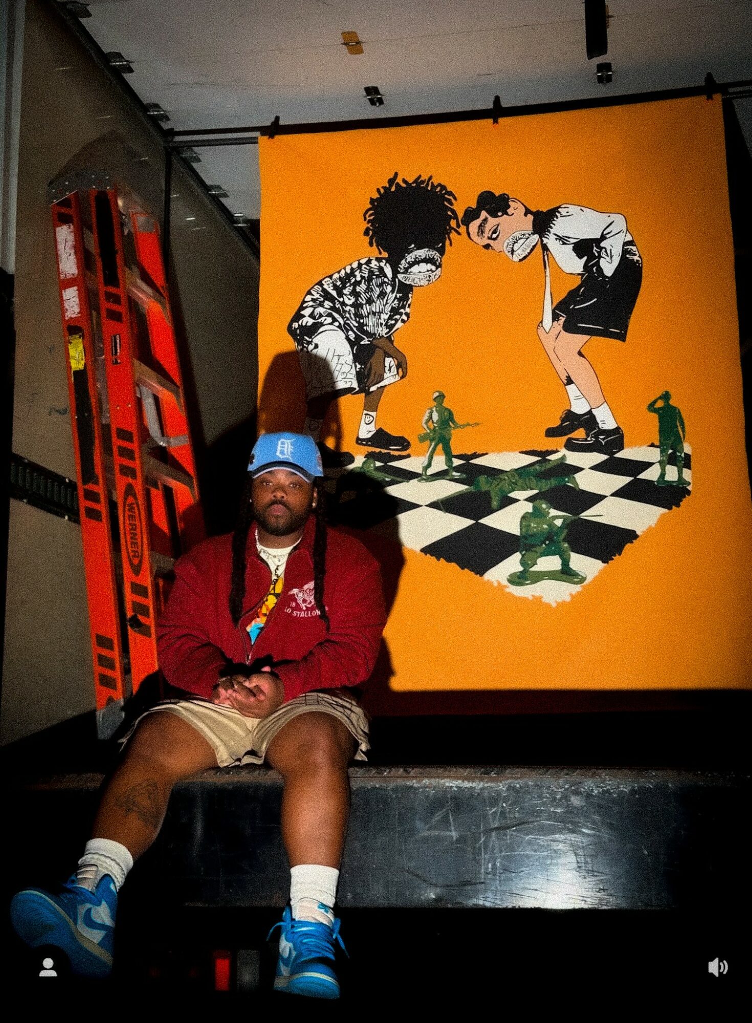 Person sitting in front of a colorful wall art with children playing chess, skis leaning against the wall, and a person wearing a blue hat.