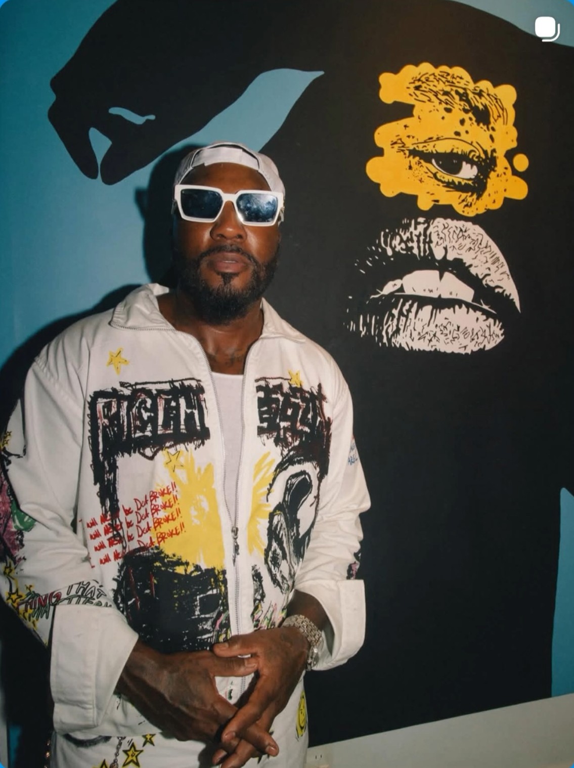 Man wearing sunglasses and a white jacket with colorful designs, standing in front of a black and yellow mural.