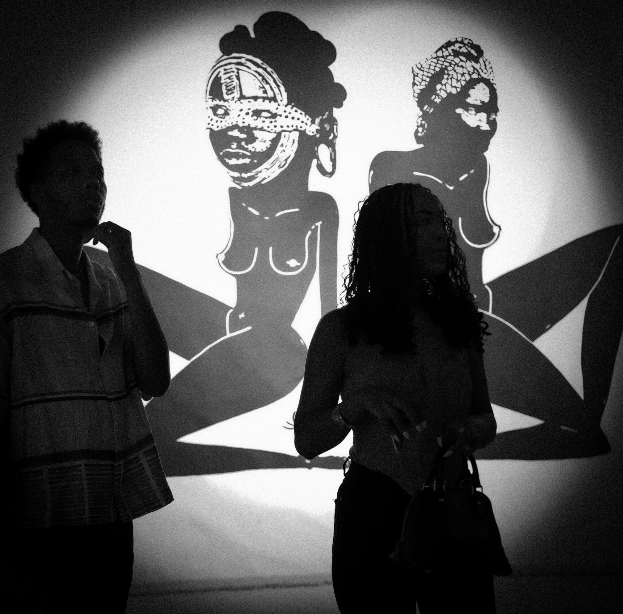 Two silhouetted people stand in front of a wall with cartoonish black and white artwork of two women.