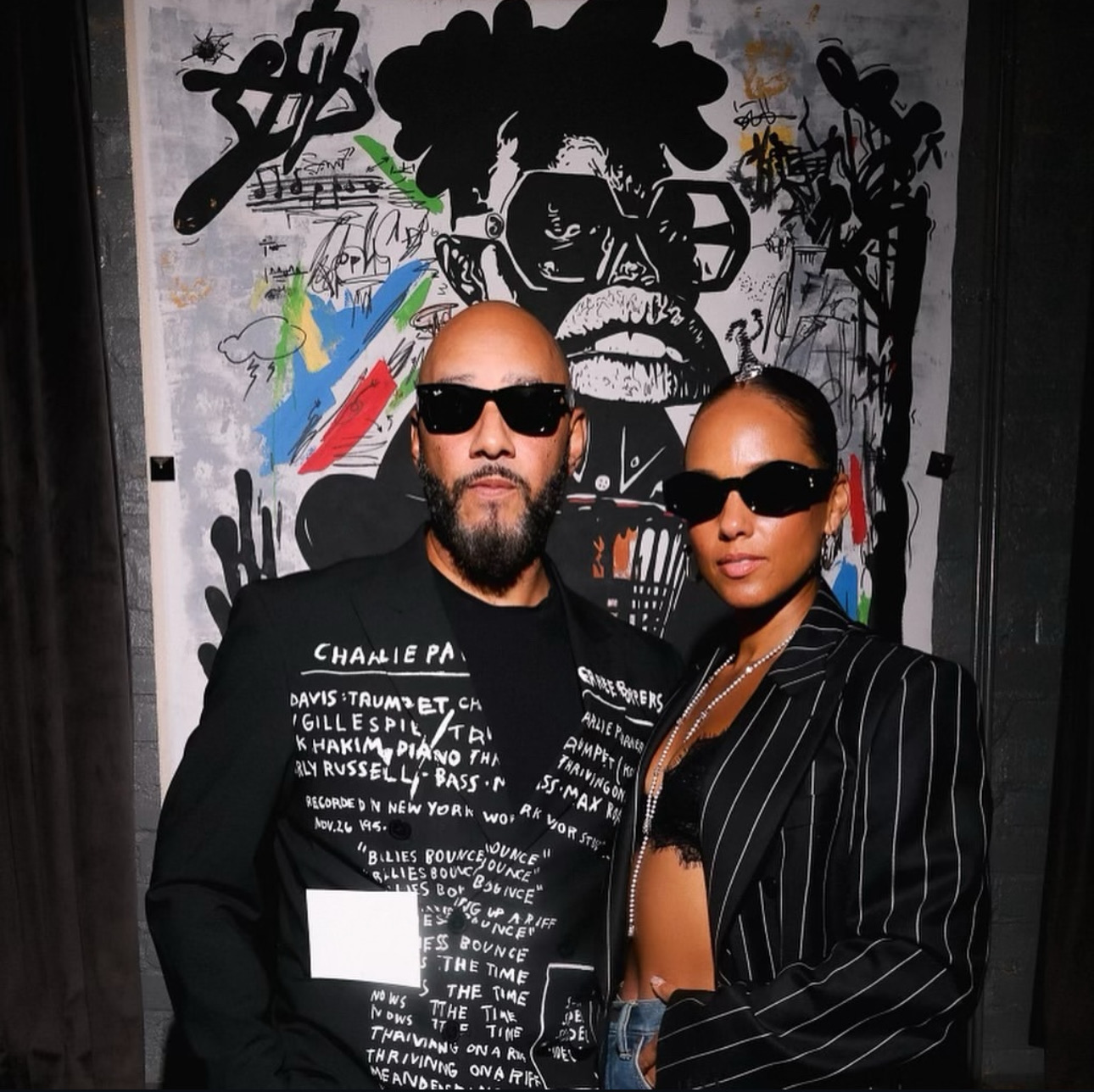 Man and woman wearing sunglasses stand in front of graffiti wall with black and white artwork.