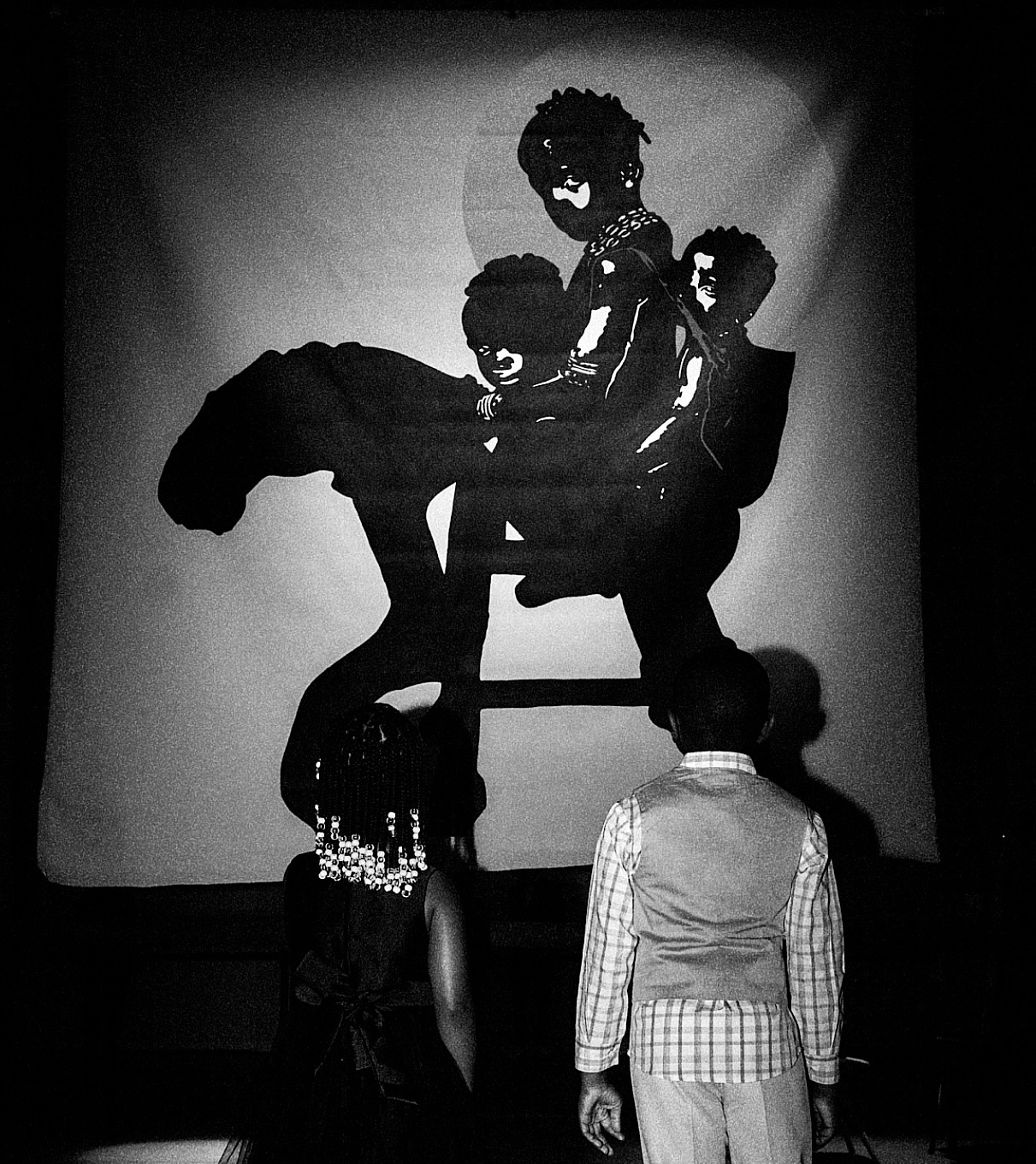 Silhouettes of three people performing a shadow puppet show on a screen, with a person watching in front.