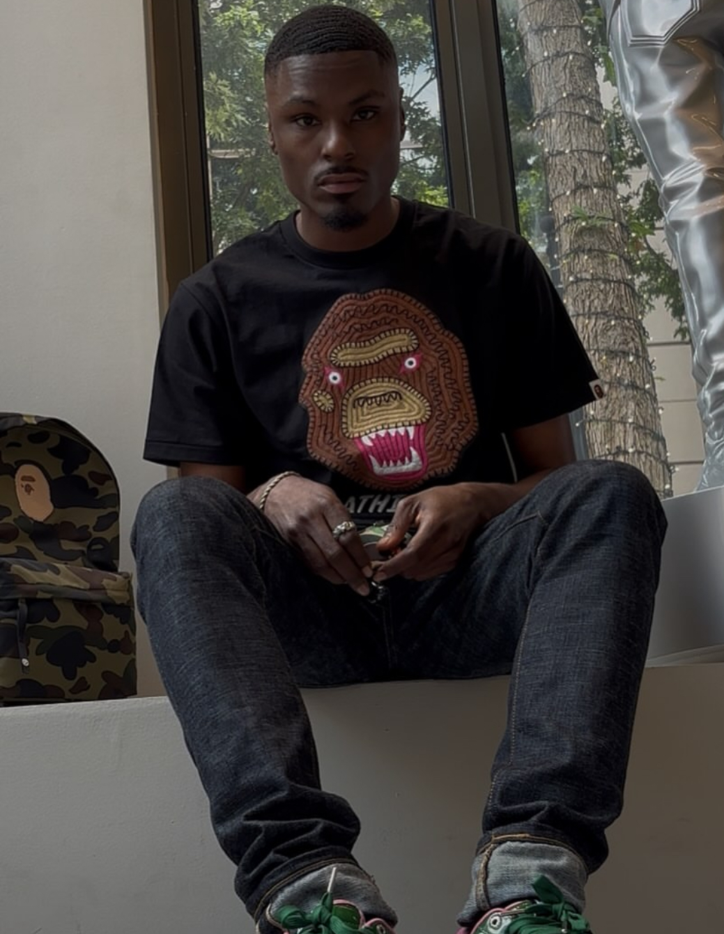 Young man sitting on a ledge indoors, wearing a black graphic t-shirt and jeans, with a window and tree outside behind him.