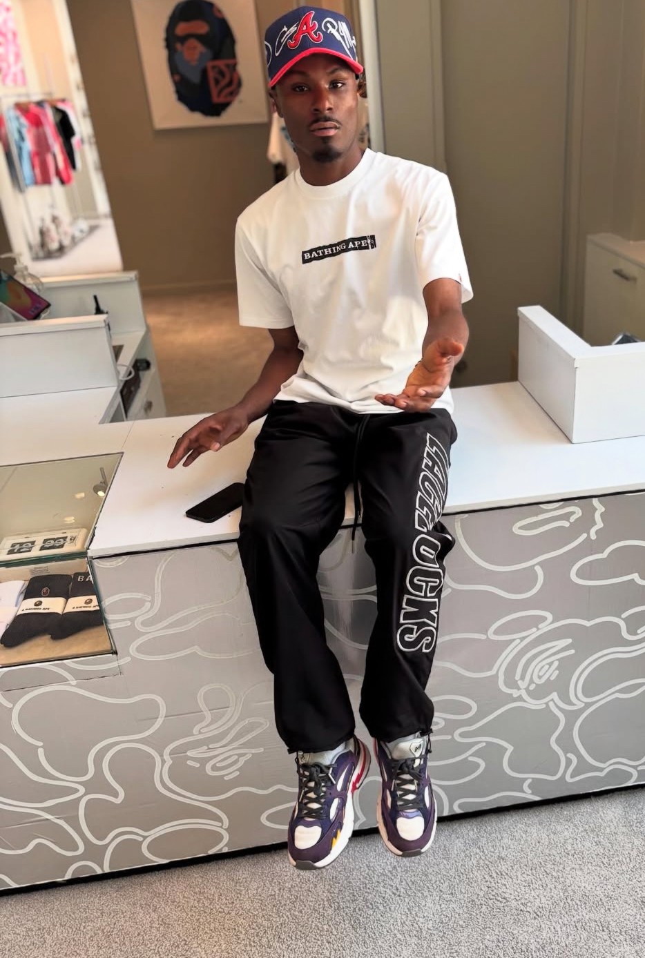 Young man sitting on a white bench in a retail store, wearing a white t-shirt, black pants, and sneakers.