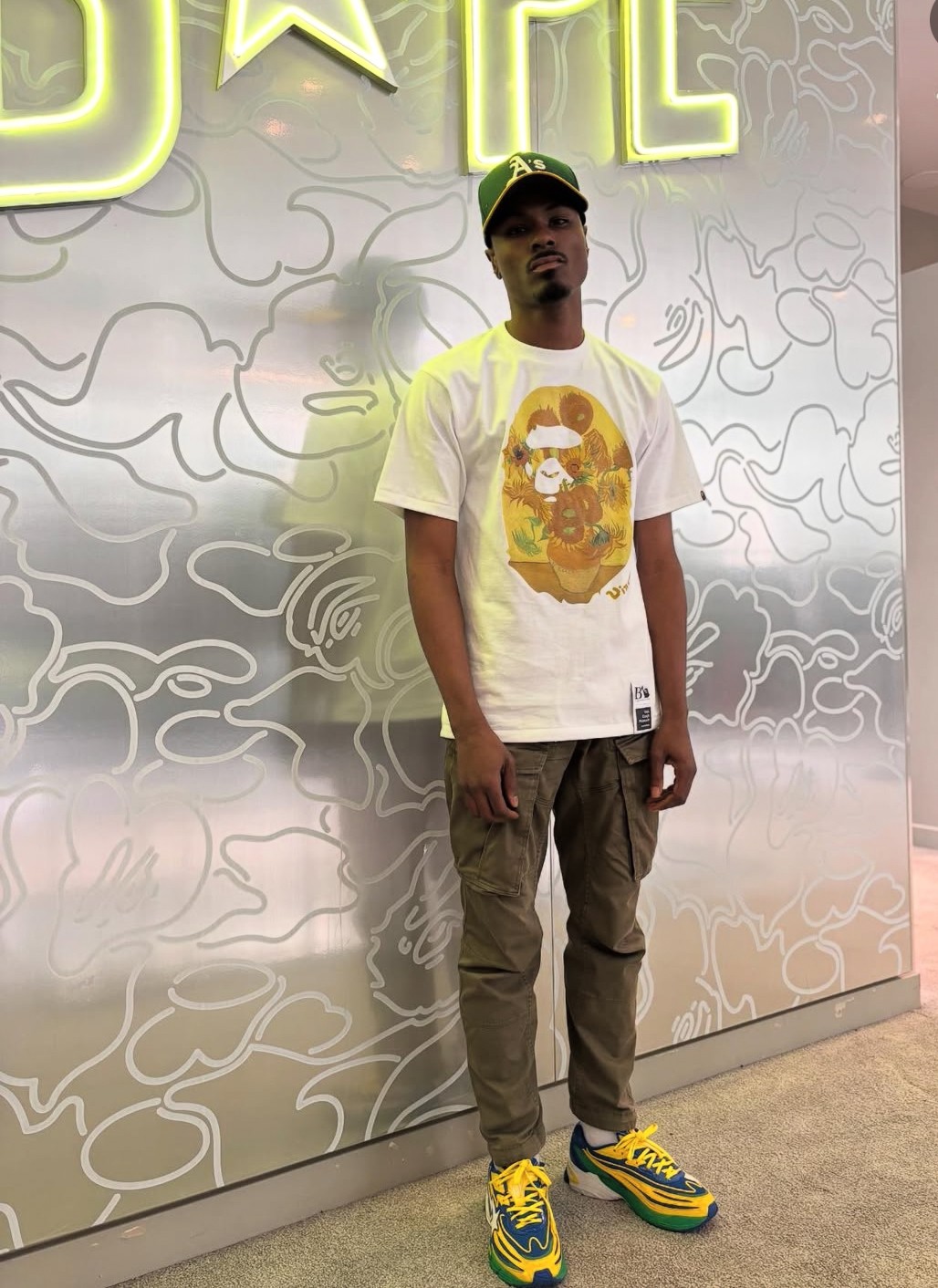 Young man standing against a patterned wall with neon sign above him, wearing a white t-shirt, cargo pants, and colorful sneakers.