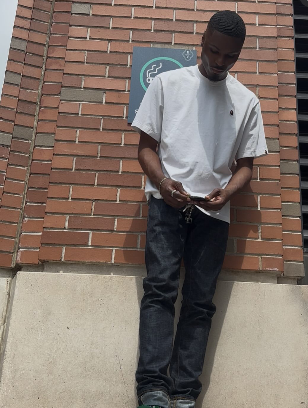 Young man standing against a brick wall, looking down at his hands, wearing a white shirt and dark jeans.