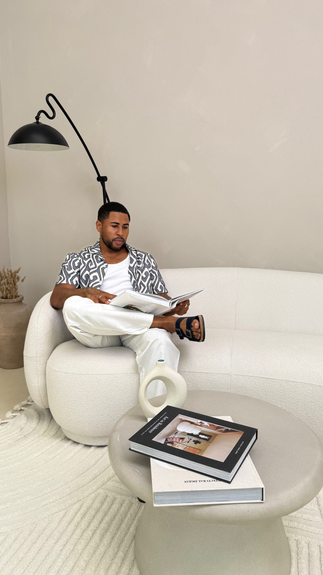 Man sitting on a white sofa reading a magazine, with a black floor lamp and a coffee table with books nearby.