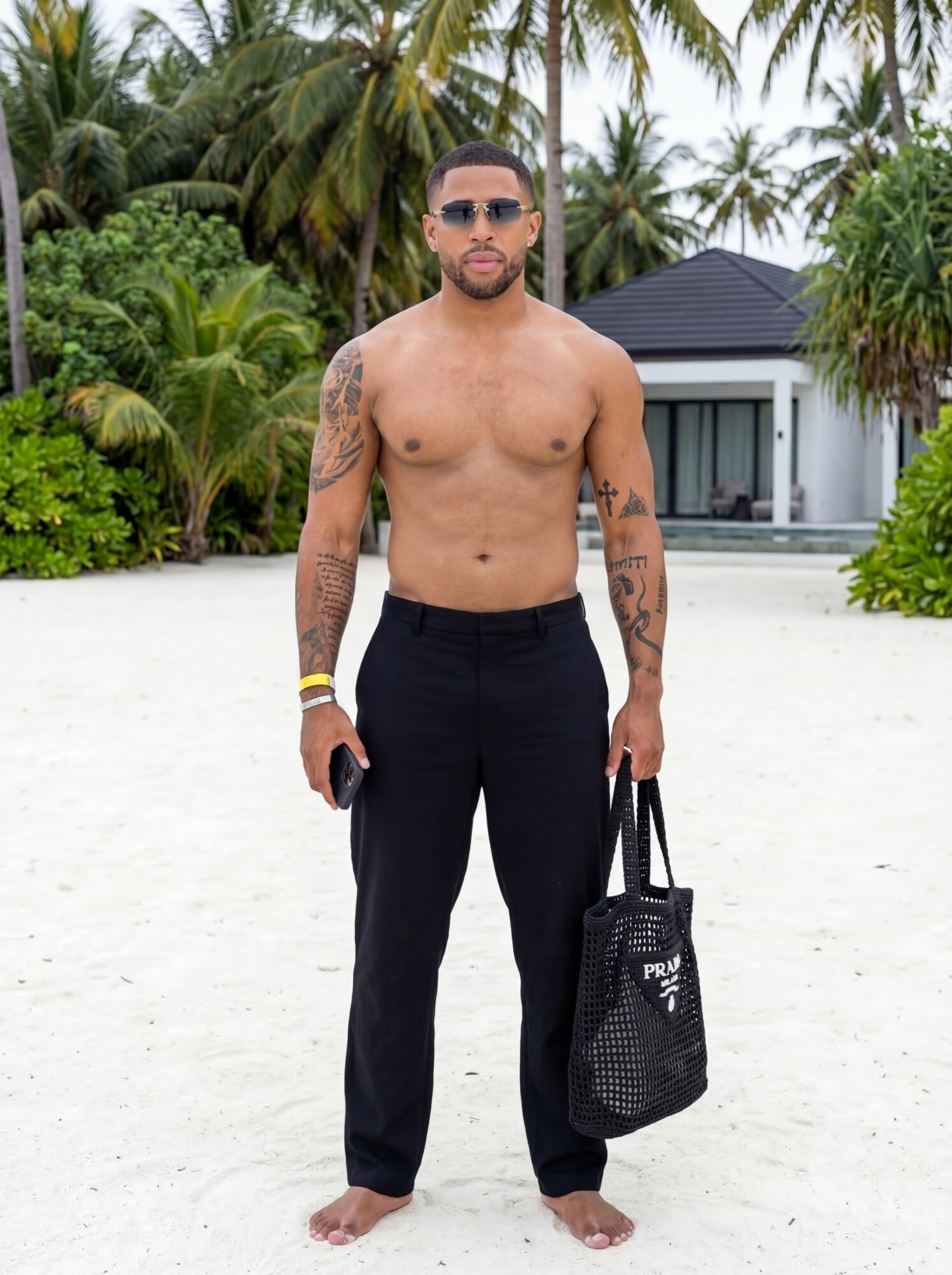 Man standing barefoot on sandy beach with palm trees and a modern house in background, holding a bag.