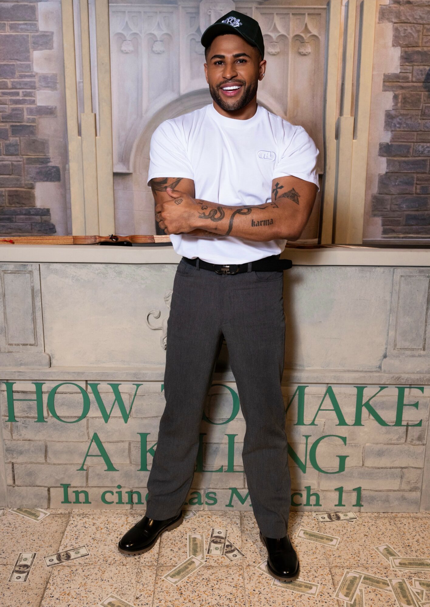 Man standing with arms crossed, smiling, wearing a white t-shirt, dark pants, and a black cap, in front of a decorative background.