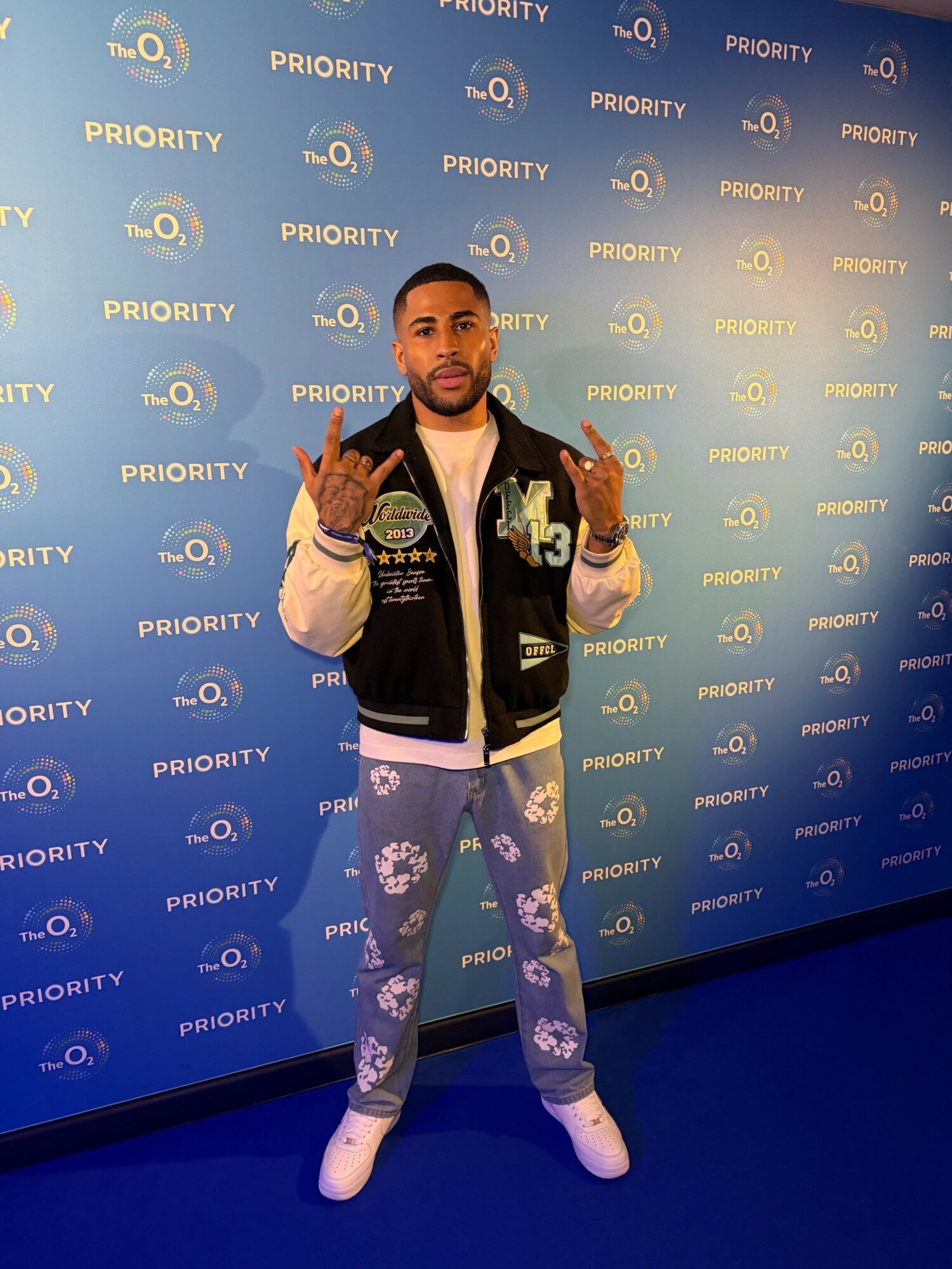 Young man standing in front of a blue backdrop with logos, making hand gestures, wearing a black jacket, white shirt, and ripped jeans.