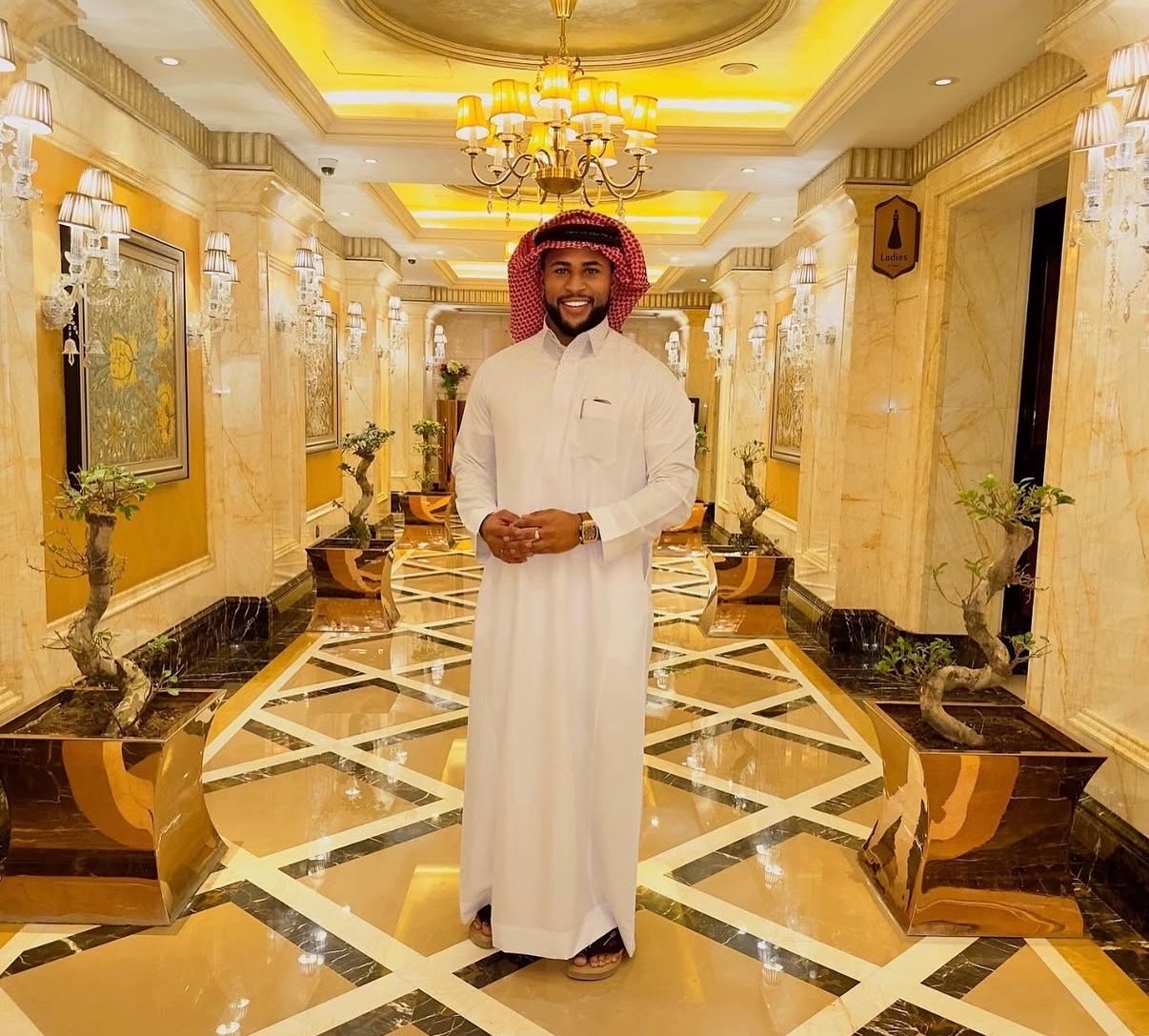 Man in traditional white attire and red head covering standing in a luxurious, ornate hallway with chandeliers and marble decor.