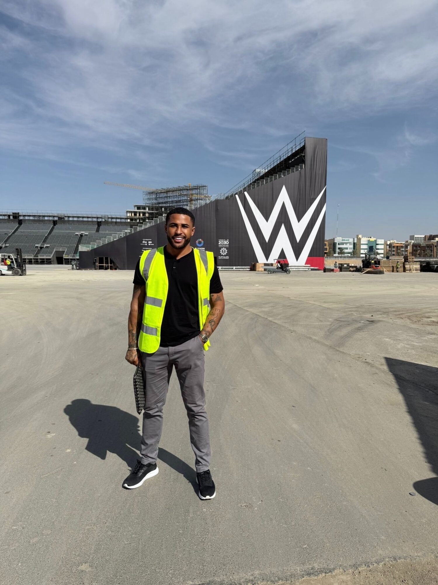 Man standing outdoors on a dirt surface, wearing a yellow safety vest, black shirt, and gray pants, with a stadium and large logo in the background.
