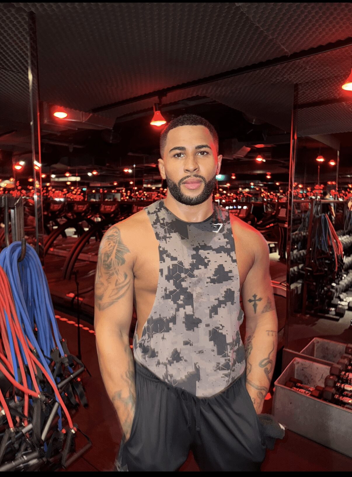 Man with tattoos wearing a sleeveless shirt, standing indoors with city lights in the background.