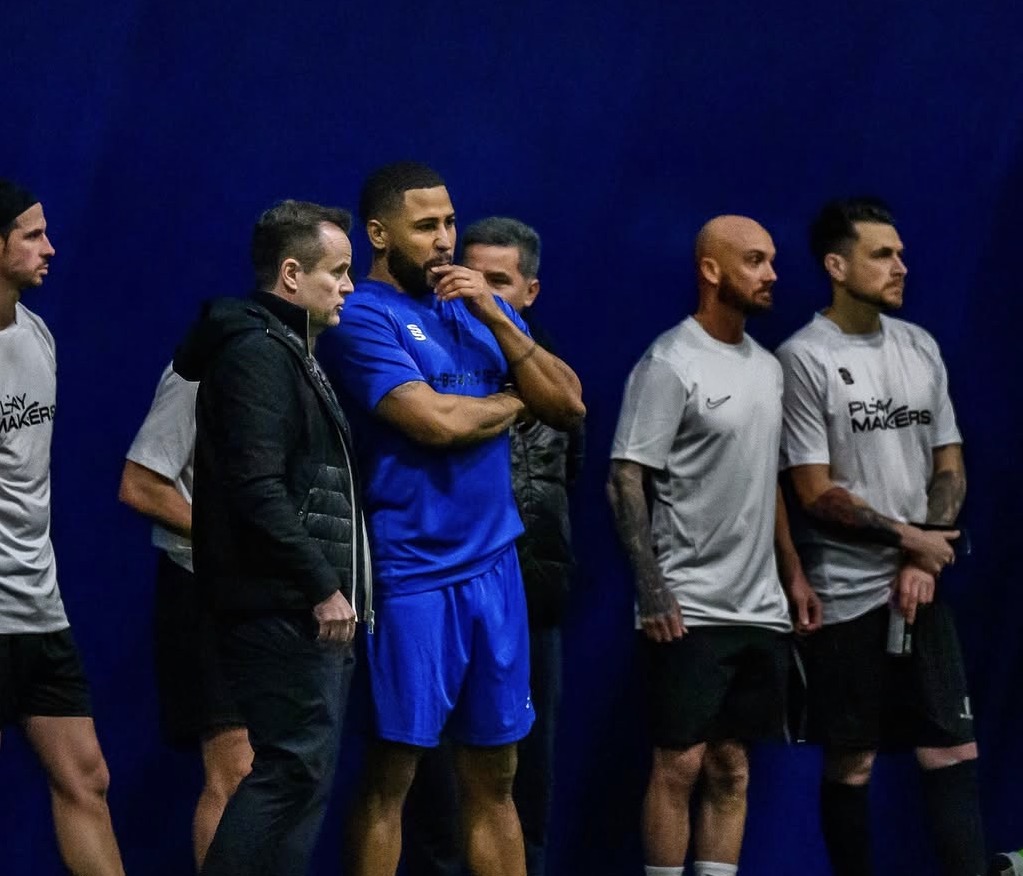 Group of six men standing against a dark blue background, some wearing sportswear, one in a blue uniform, others in casual clothing.