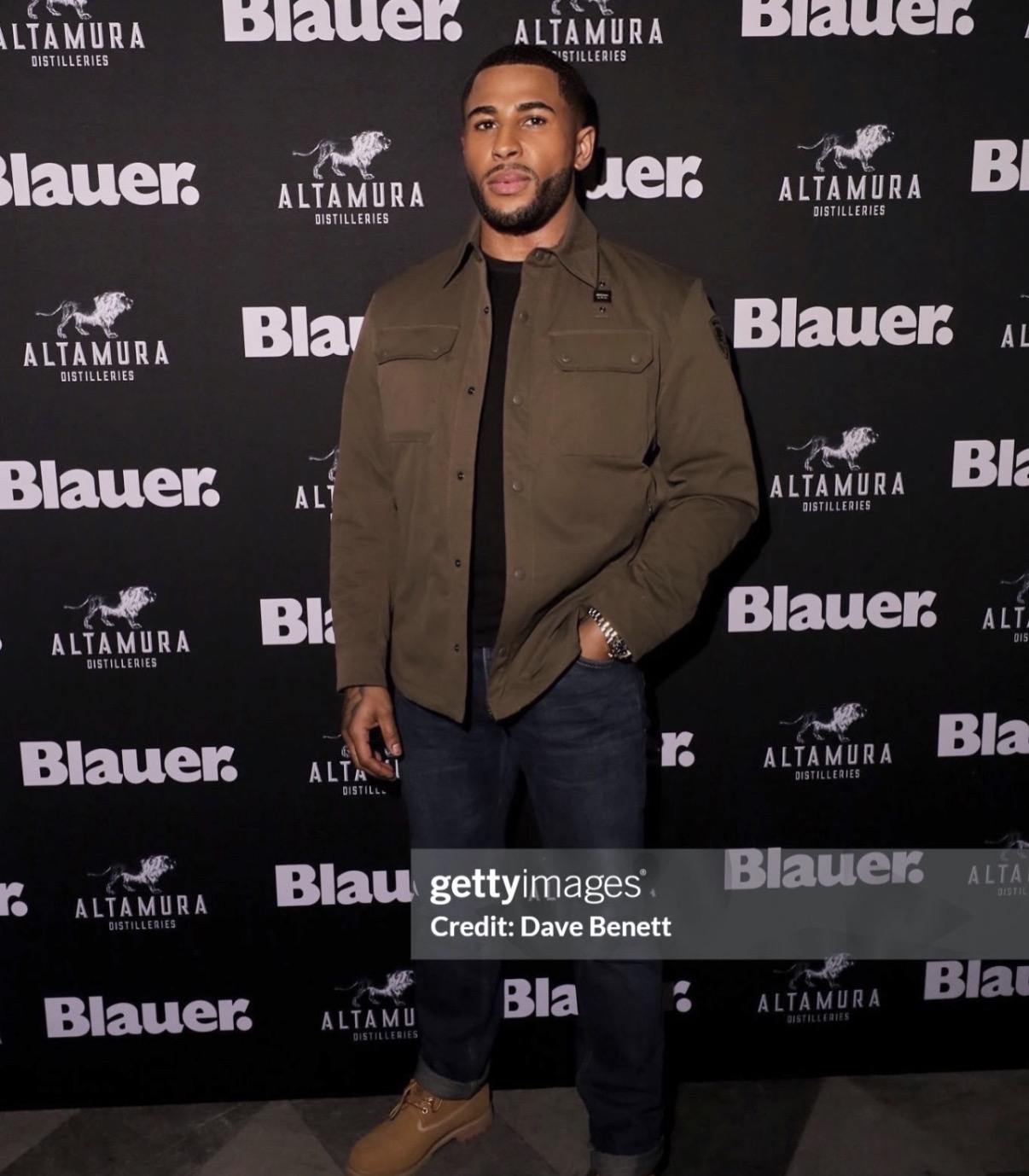 Man standing in front of a black backdrop with white logos, wearing a brown jacket and dark pants.