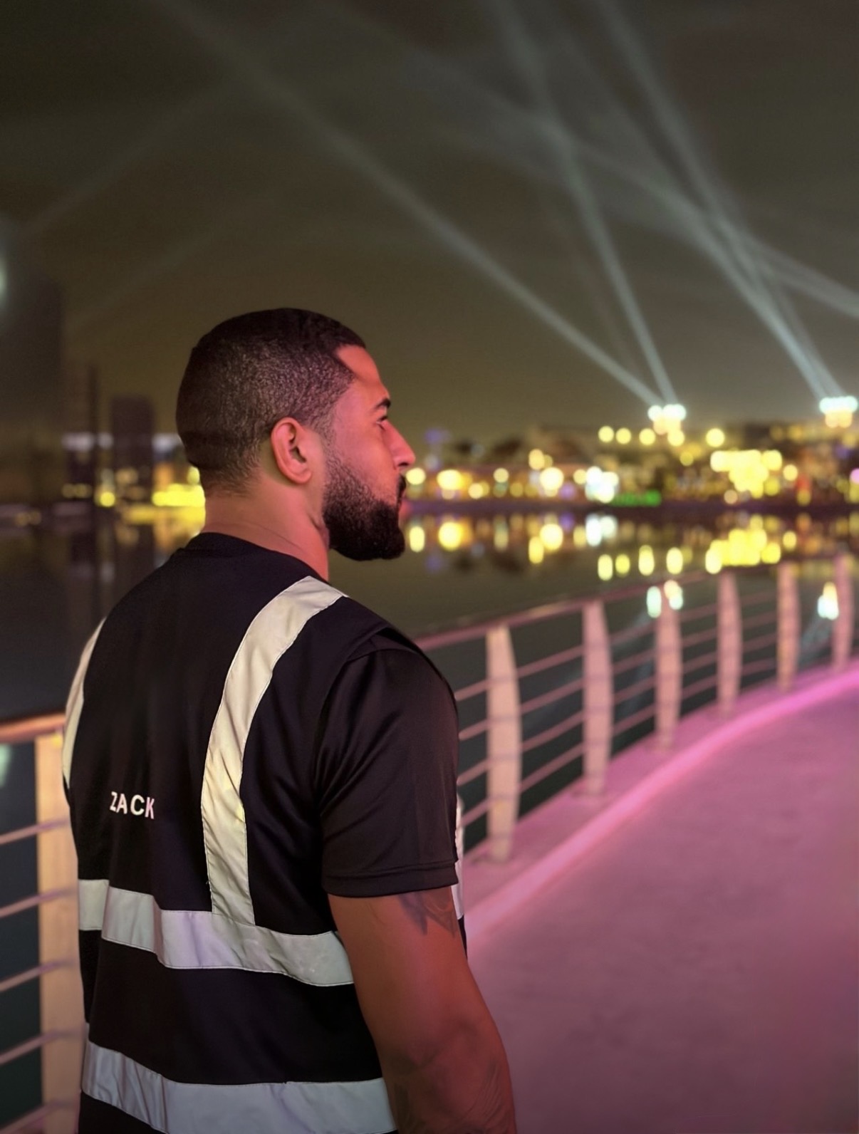 Side view of a man with short hair and beard standing on a bridge at night with city lights and light beams in background.