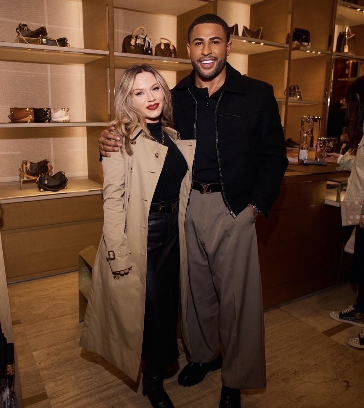 Two people standing together in a store, shelves with handbags behind them, smiling at the camera.