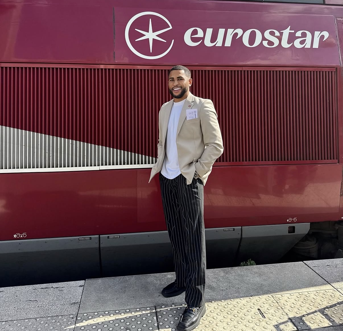 Man standing in front of a red Eurostar train, smiling, wearing a beige blazer and striped pants.
