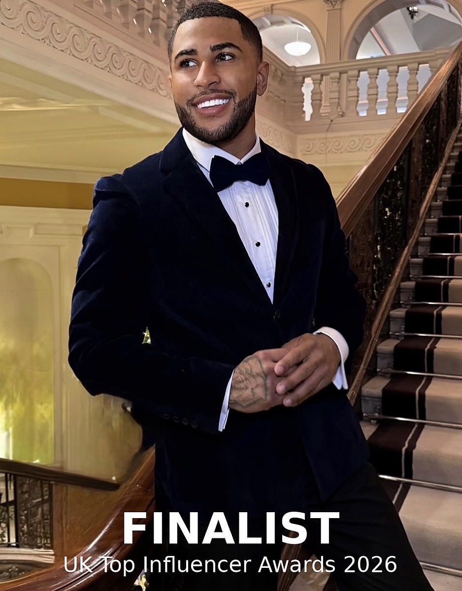Man in tuxedo standing on staircase inside elegant building, smiling, with ornate architecture in background.