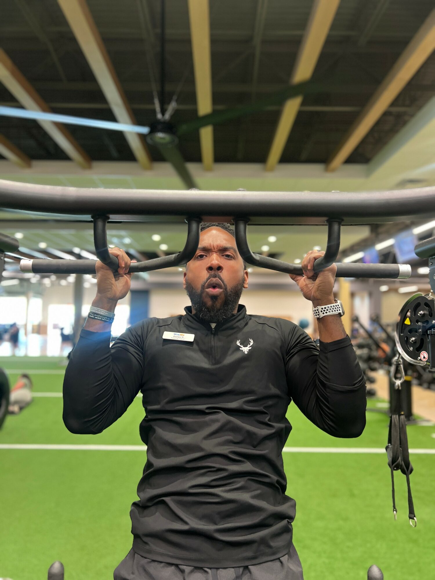 Man lifting a barbell overhead in a gym with green flooring and ceiling fan.
