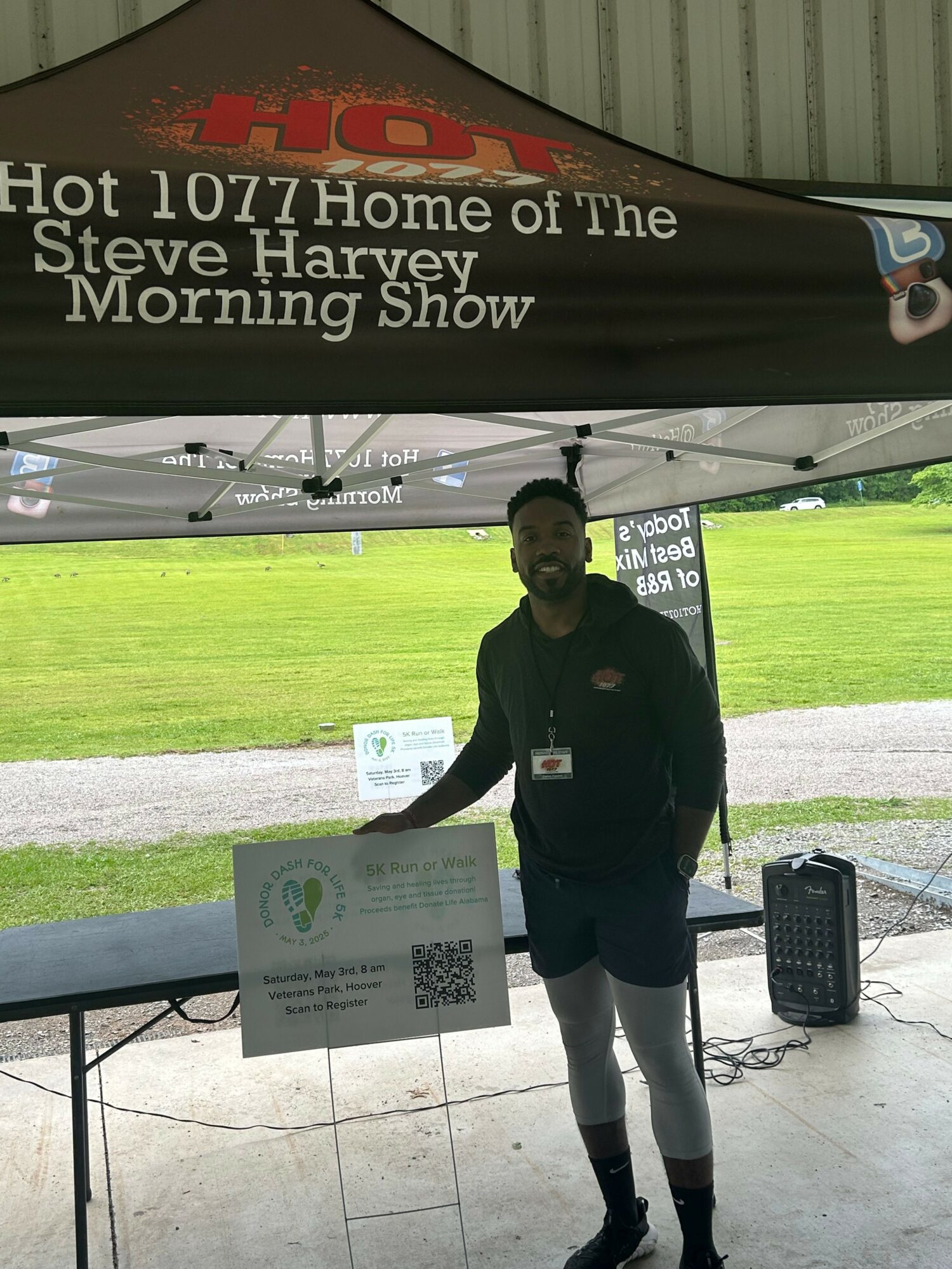 Person standing under a tent with a sign, outdoors with green grass background, holding a paper, wearing dark clothing and shorts.
