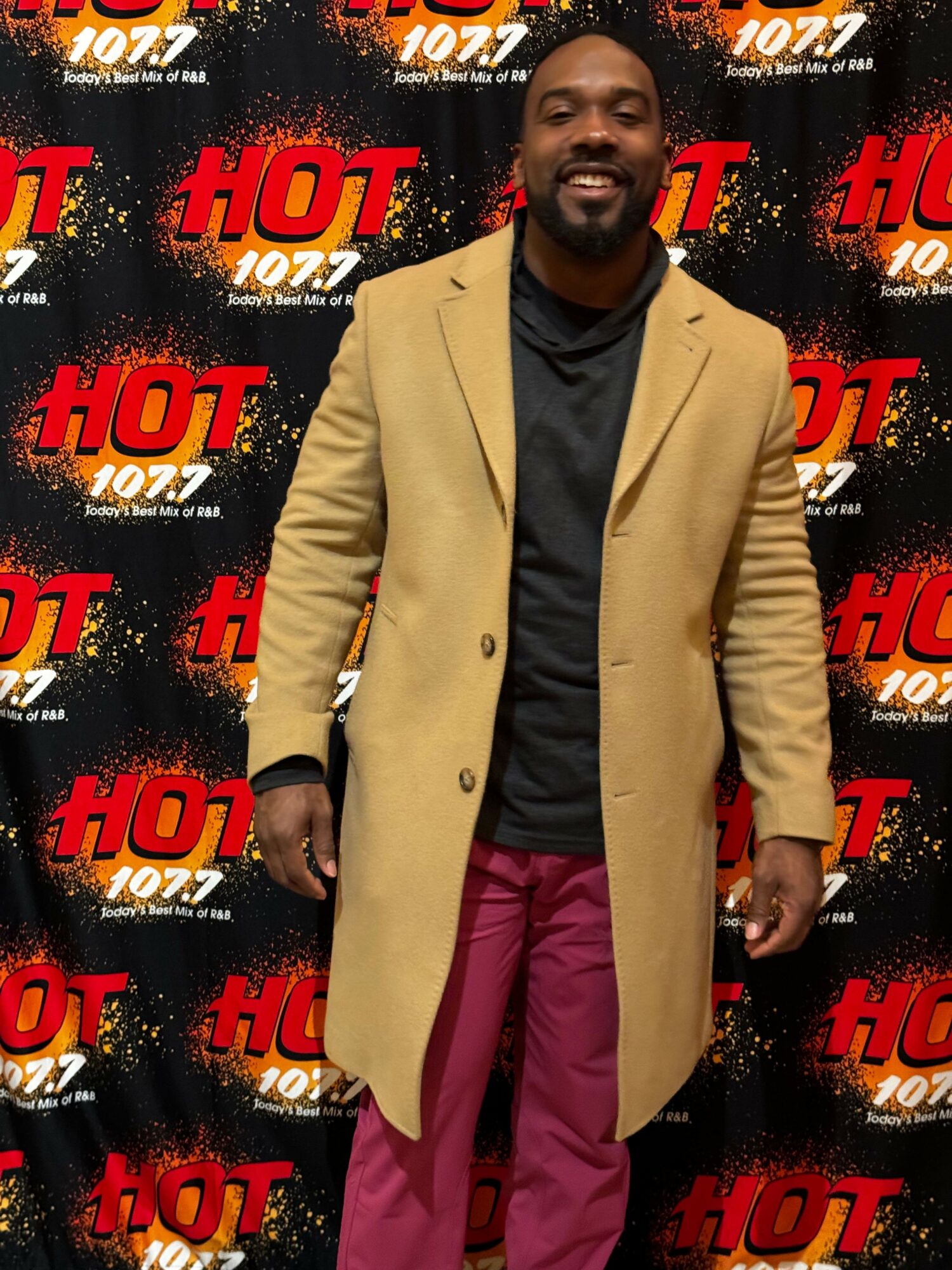 Man in beige coat and pink pants standing in front of a black background with red and white 'HOT 107.7' logos.
