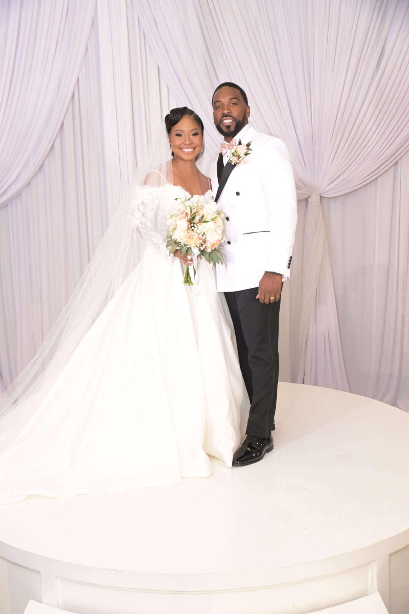 Bride and groom standing together on stage with white curtain backdrop, bride holding a bouquet, both smiling.