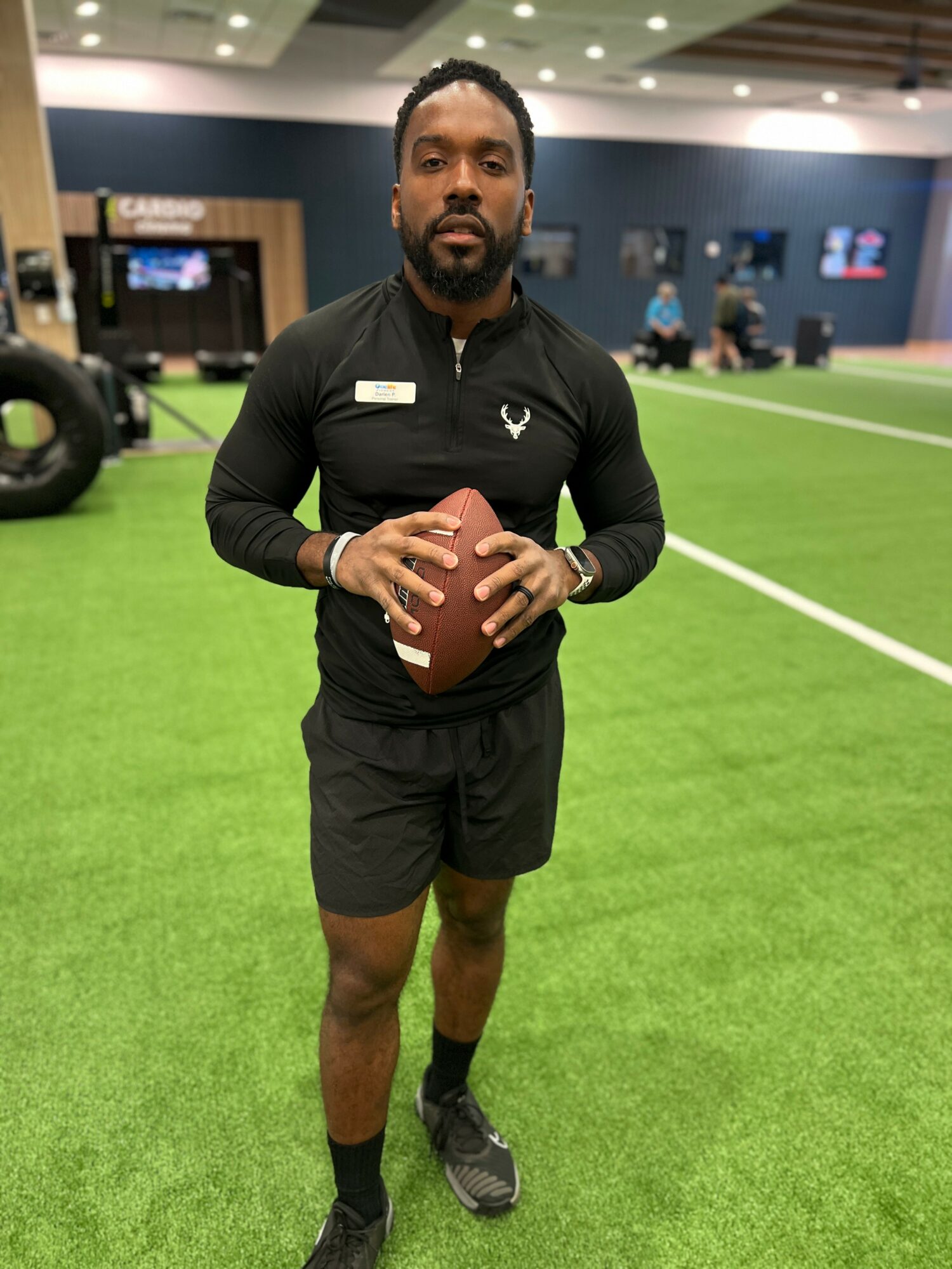 Man holding football on indoor field with green turf and blue walls, wearing black sportswear and sneakers.