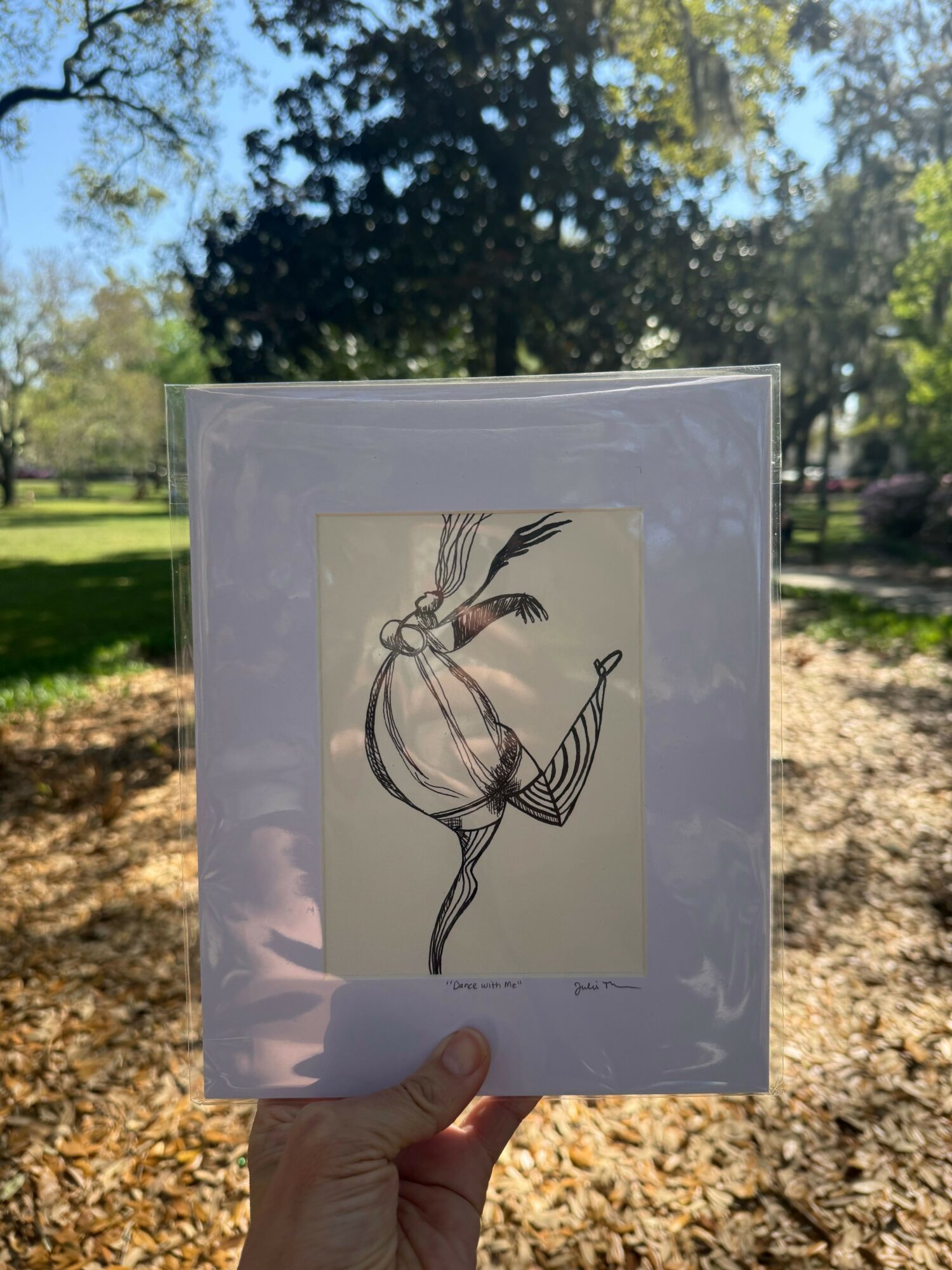 Person holding a framed artwork of a stylized bird with long feathers outdoors on a sunny day.
