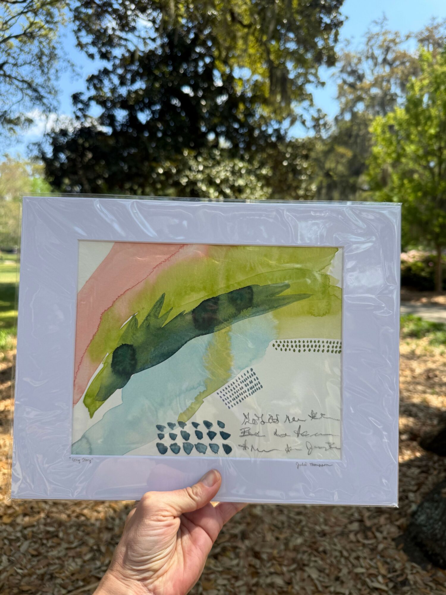 Hand holding a framed abstract watercolor painting outdoors with trees and sky in background.