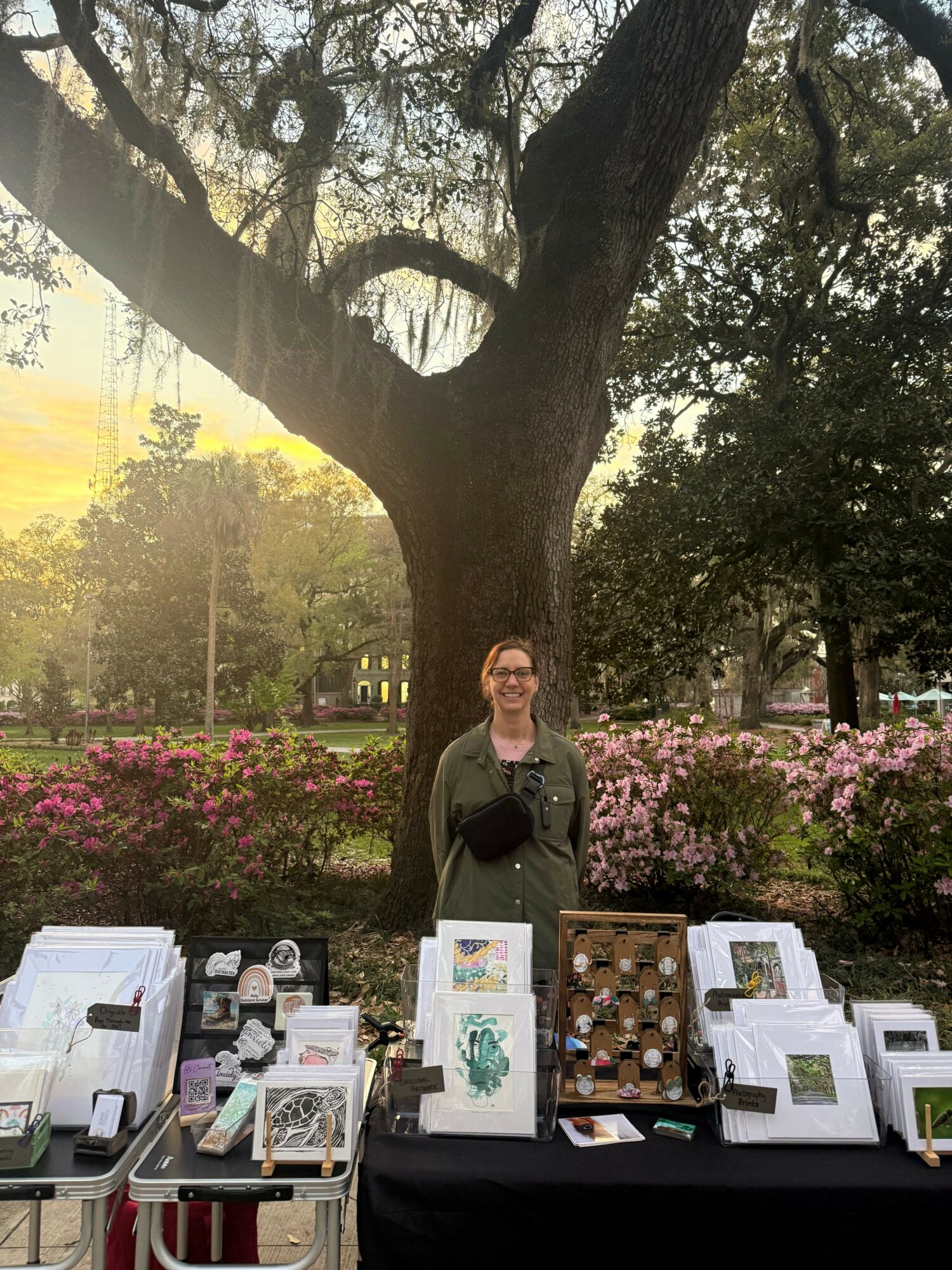 Woman standing behind a table with artwork and jewelry outdoors, large tree and pink flowers in background.