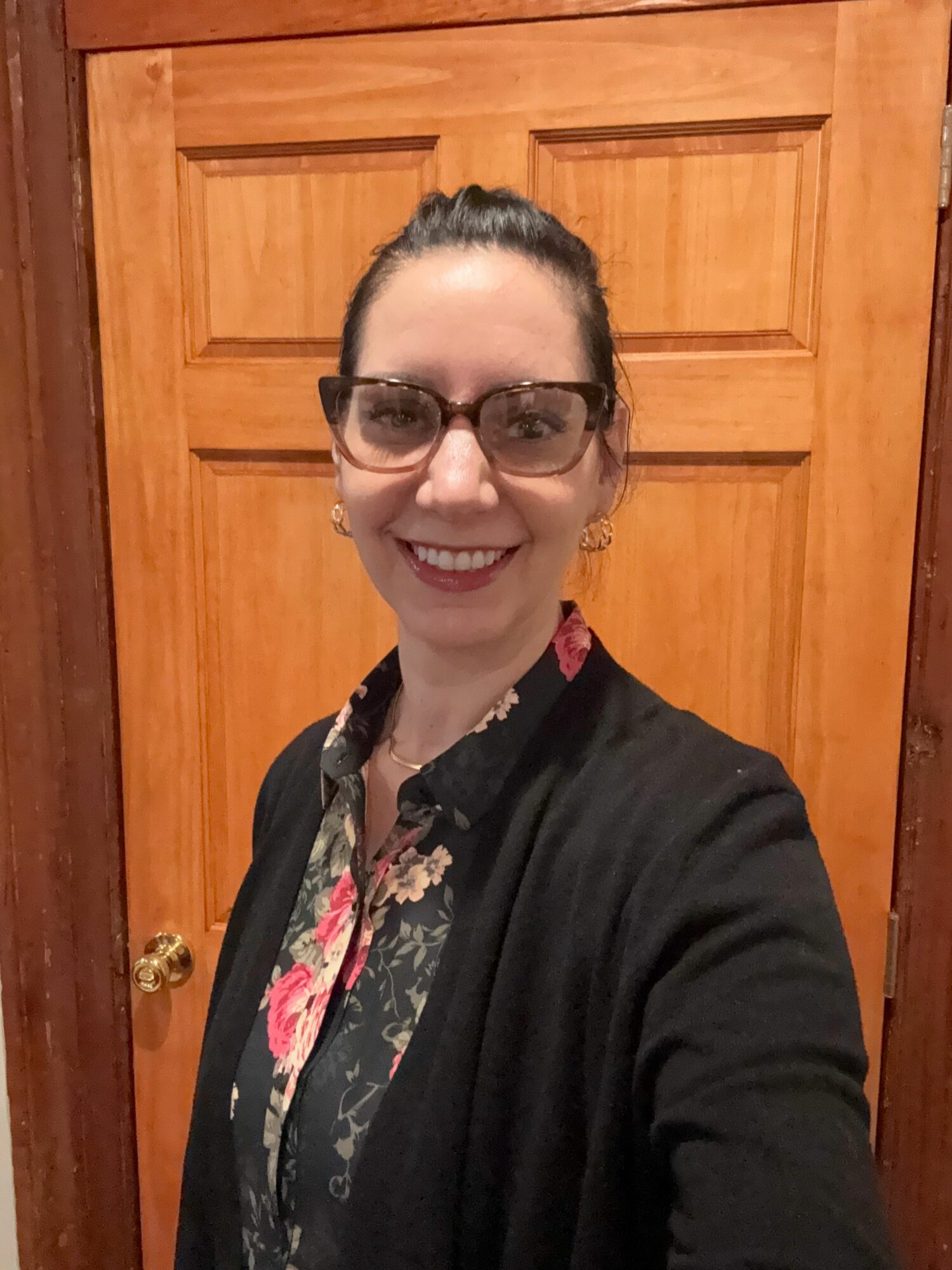 Woman with glasses smiling, wearing a floral shirt and black cardigan, standing in front of a wooden door.