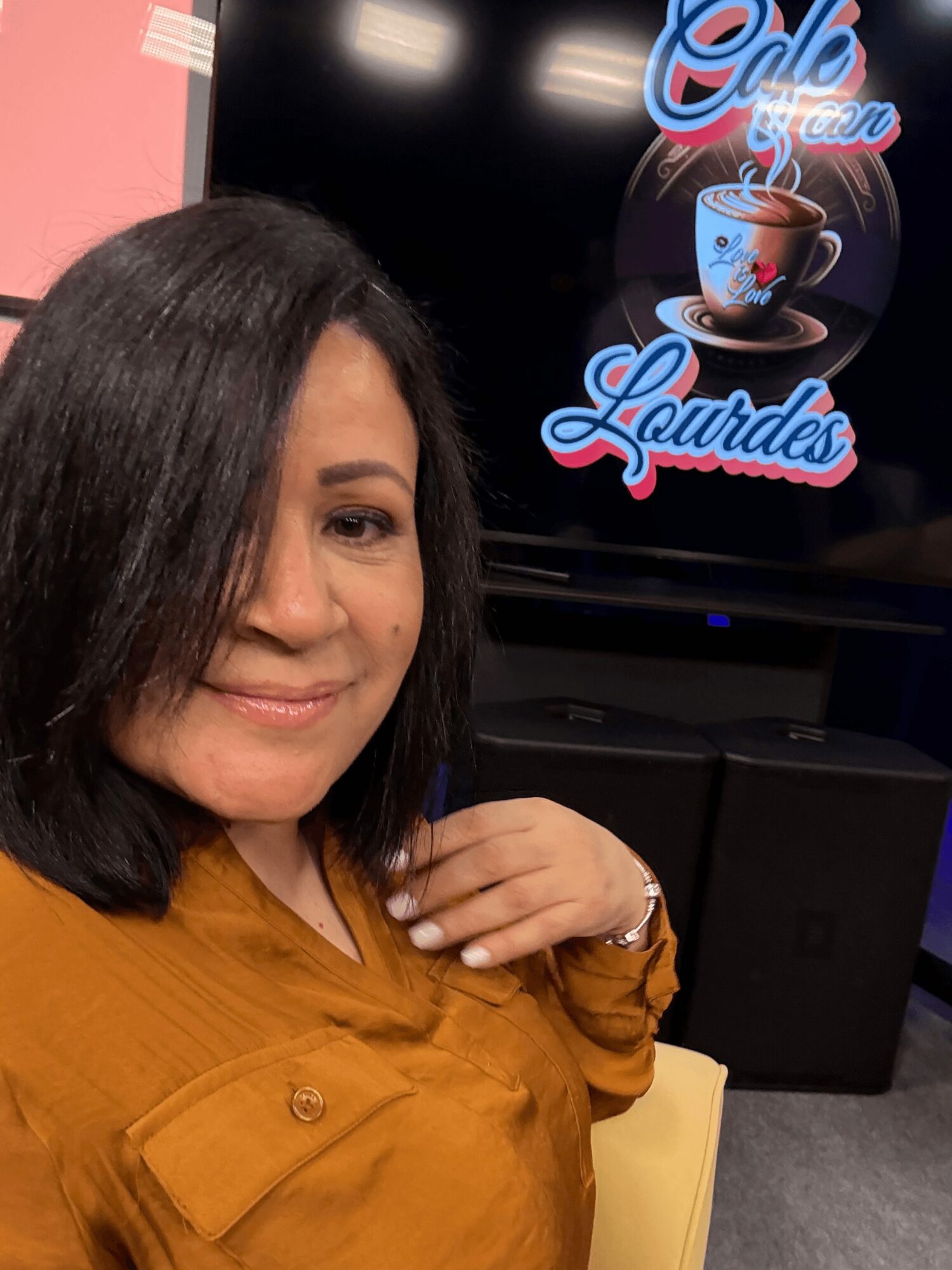 Woman with shoulder-length dark hair smiling, wearing a mustard-colored top, in front of a screen with a coffee cup logo.
