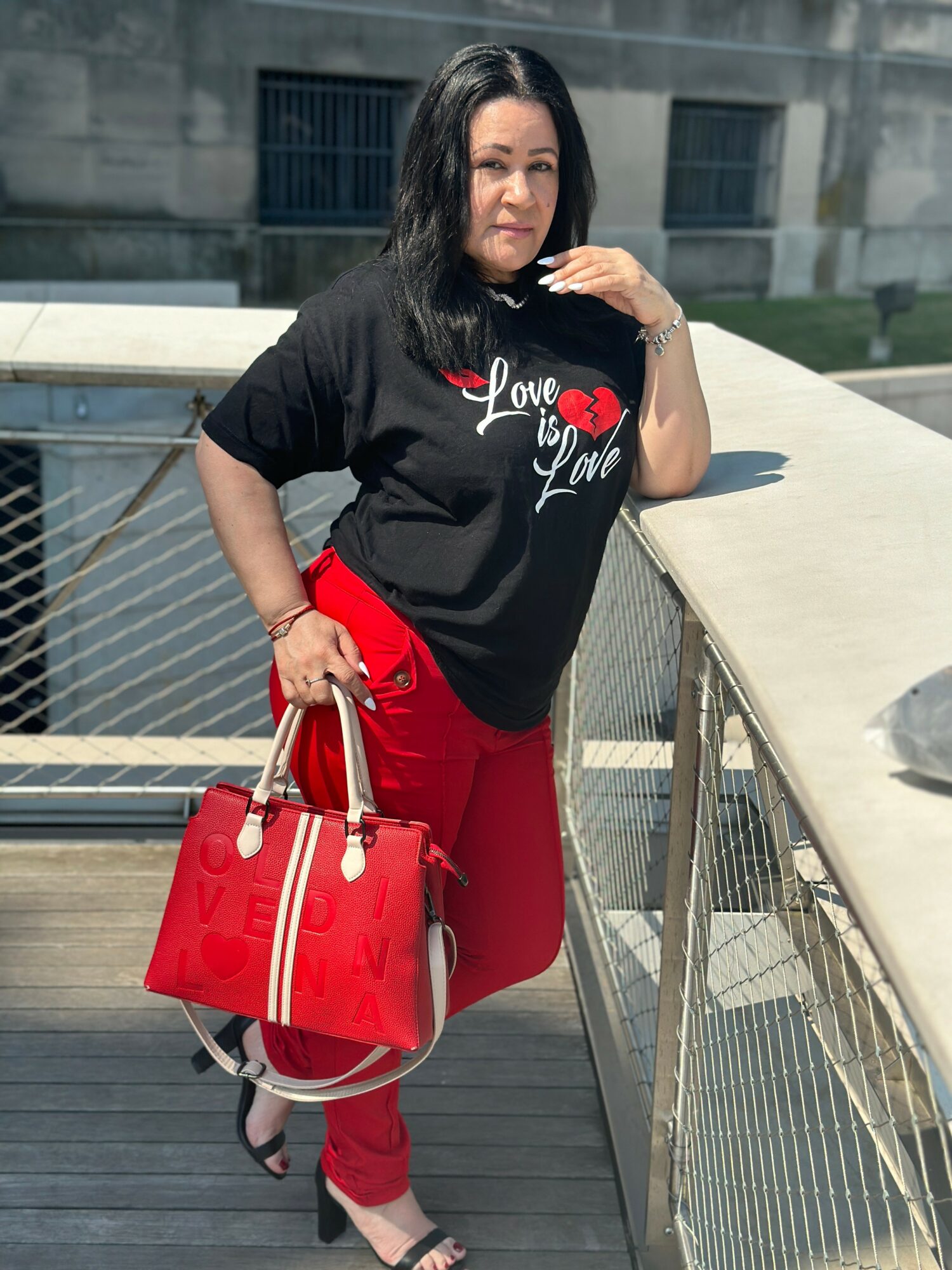 Woman standing on a balcony with a railing, wearing a black shirt with red and white text, red pants, and holding a red handbag.