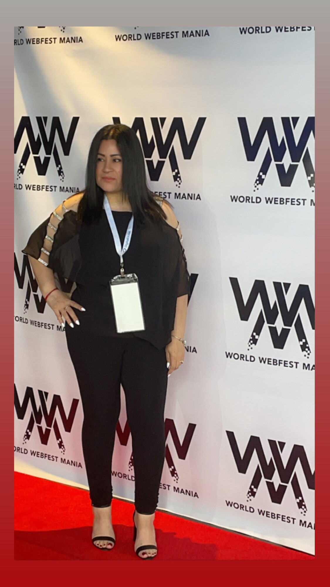 Woman standing on red carpet in front of backdrop with logos, wearing black outfit and lanyard.
