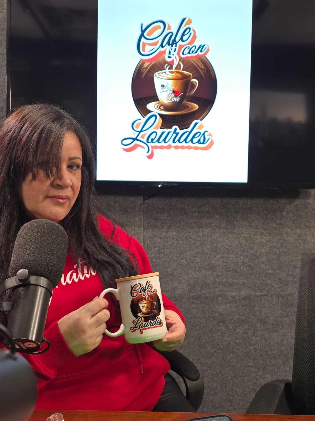 Woman in red sweater holding a coffee mug with a logo, sitting in front of a sign that says 