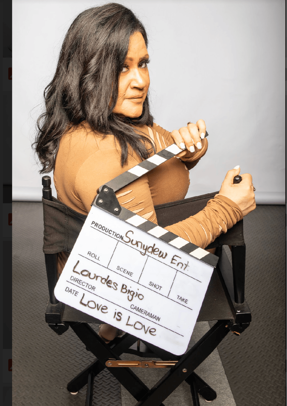 Woman with dark wavy hair sitting on a chair, holding a clapperboard, wearing a brown long-sleeve top.