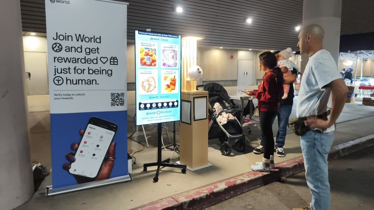 People standing near digital screens and a banner at night outside a building, with strollers nearby.