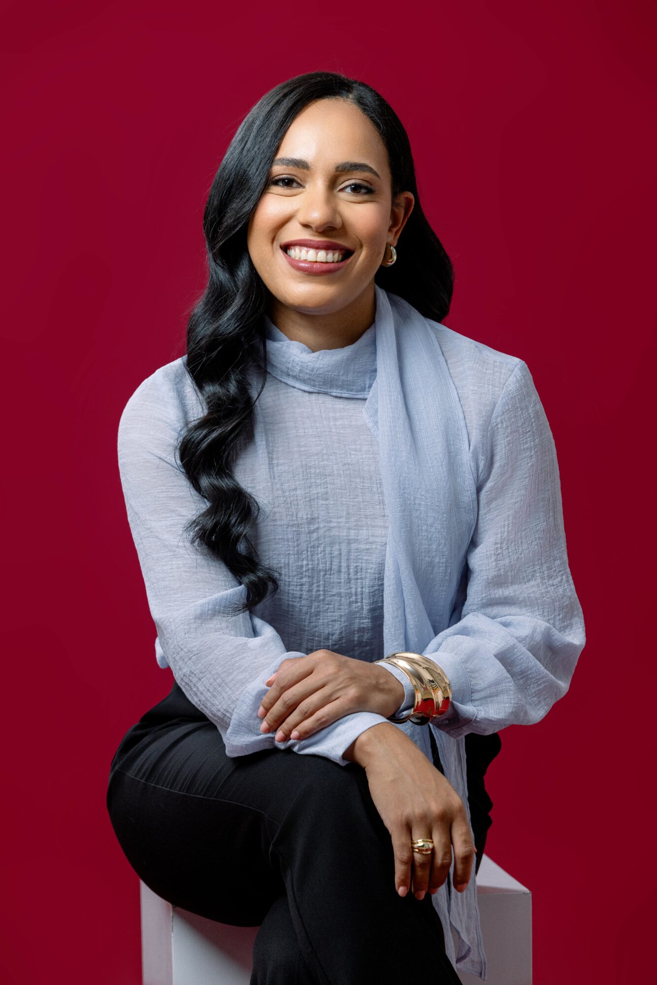 Smiling woman with long black hair, wearing a light blue sweater and scarf, sitting against a red background.