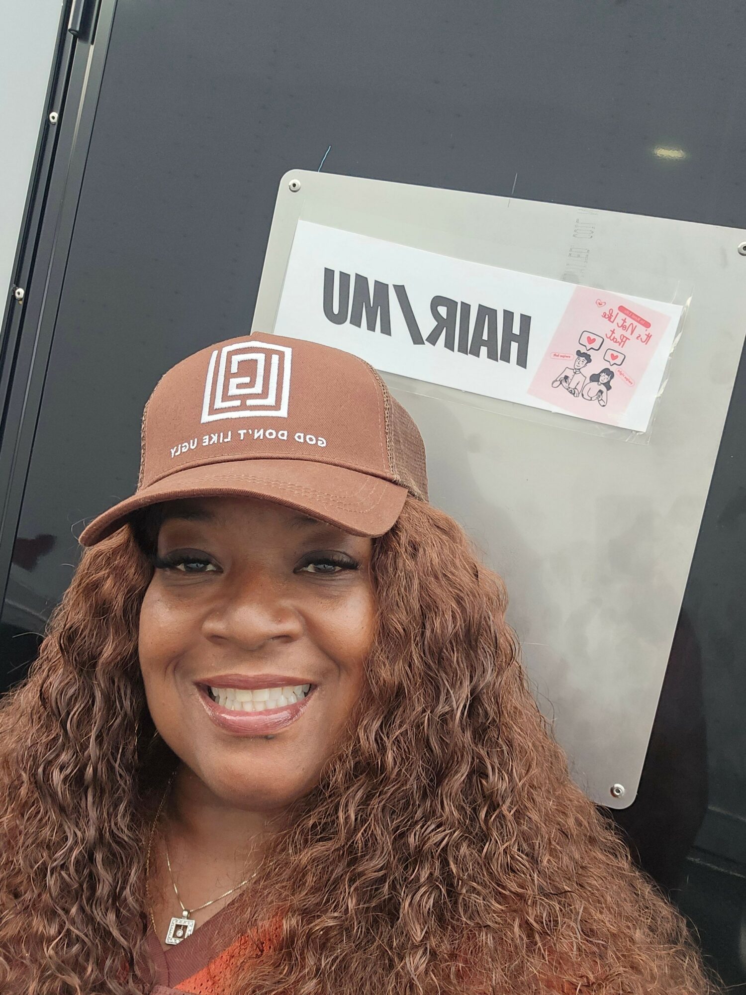 Woman with curly hair wearing a brown cap smiling in front of a sign with text.