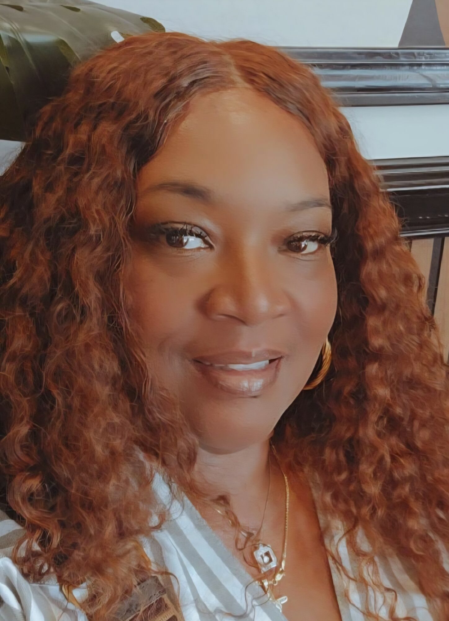 Woman with curly reddish-brown hair, wearing a striped shirt and gold jewelry, smiling at camera.
