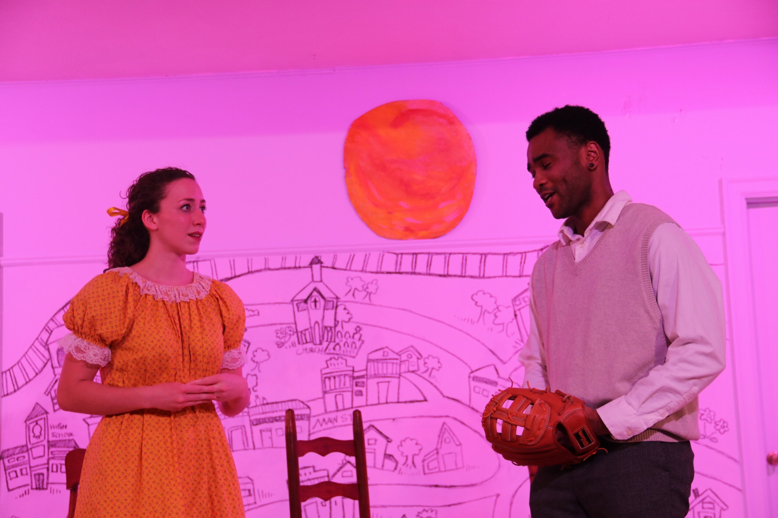 Two people standing in front of a pink wall with a painted sun and landscape, engaging in conversation.