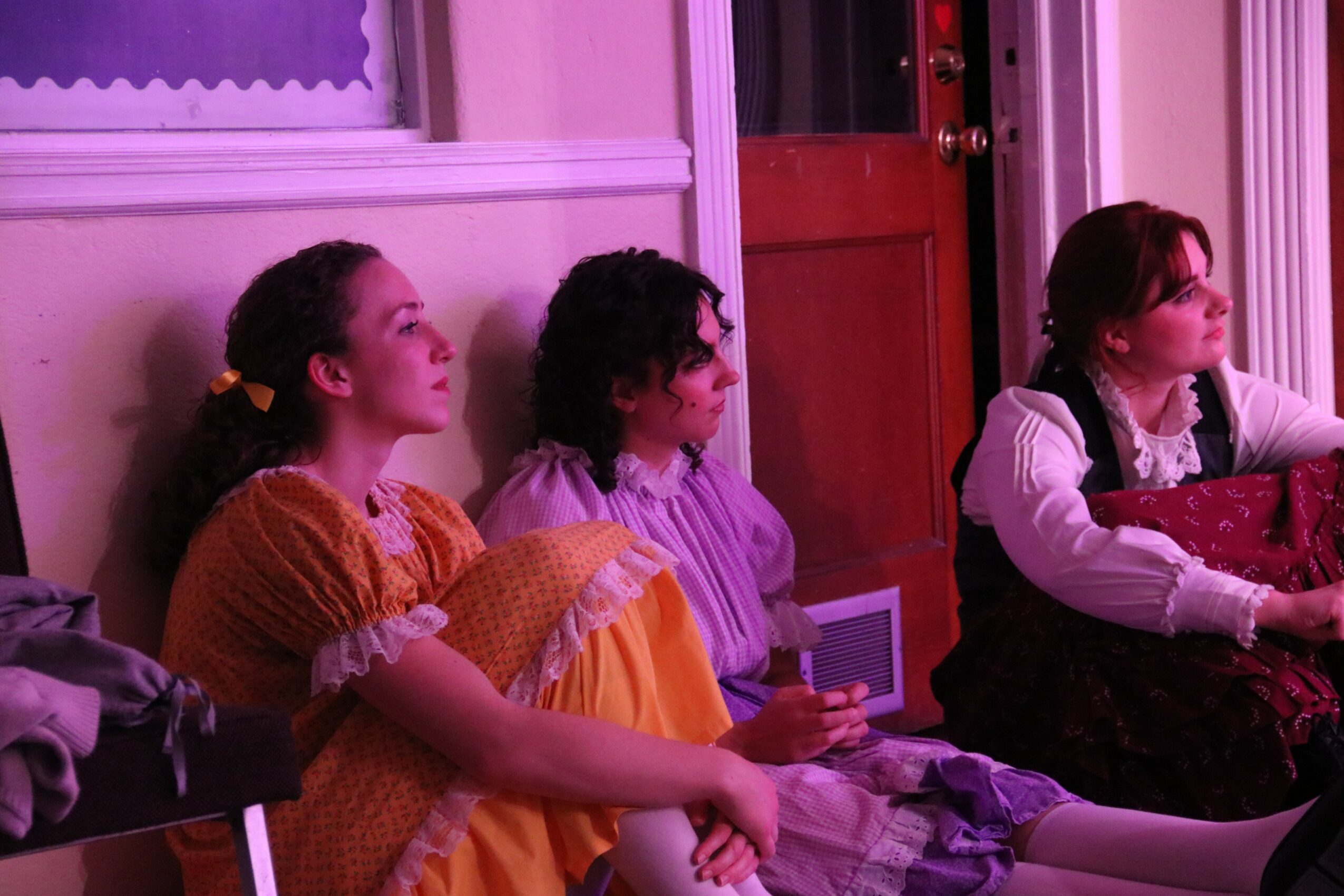 Three women sitting on a bench indoors, facing right, with a door behind them and pink lighting.