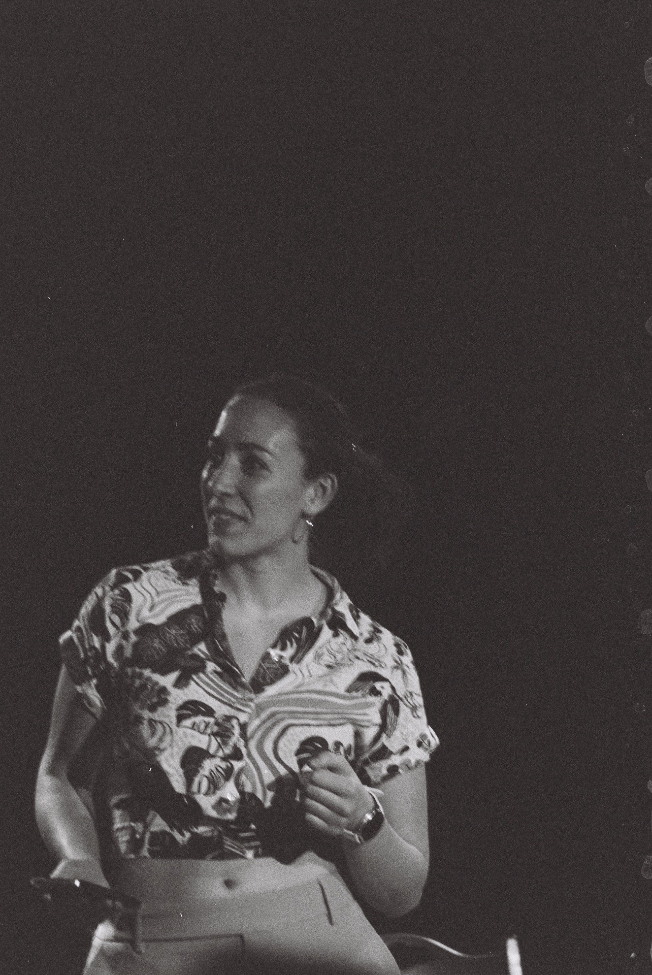 Woman holding a microphone, smiling, wearing a patterned shirt, standing against a dark background.