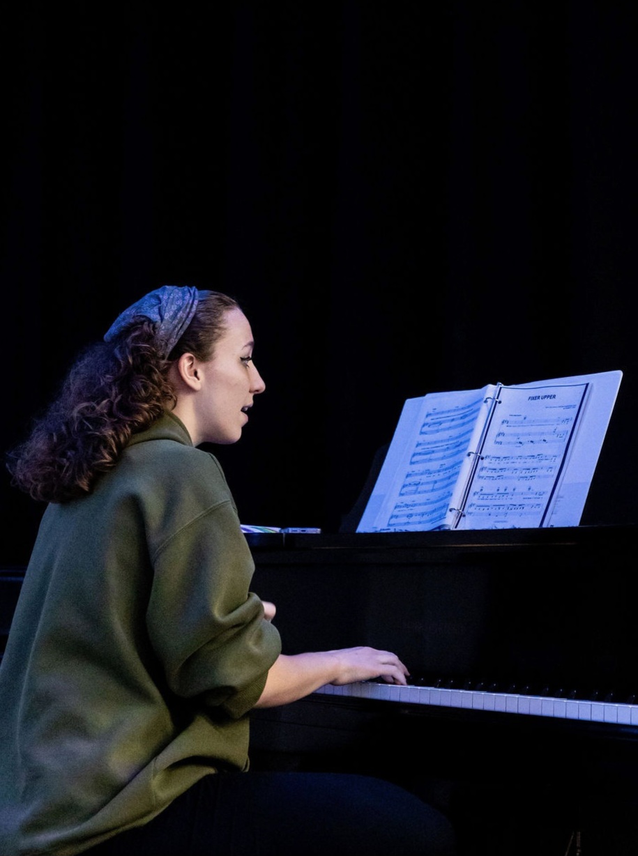 Woman playing piano with sheet music on stand, dark background, side profile view.