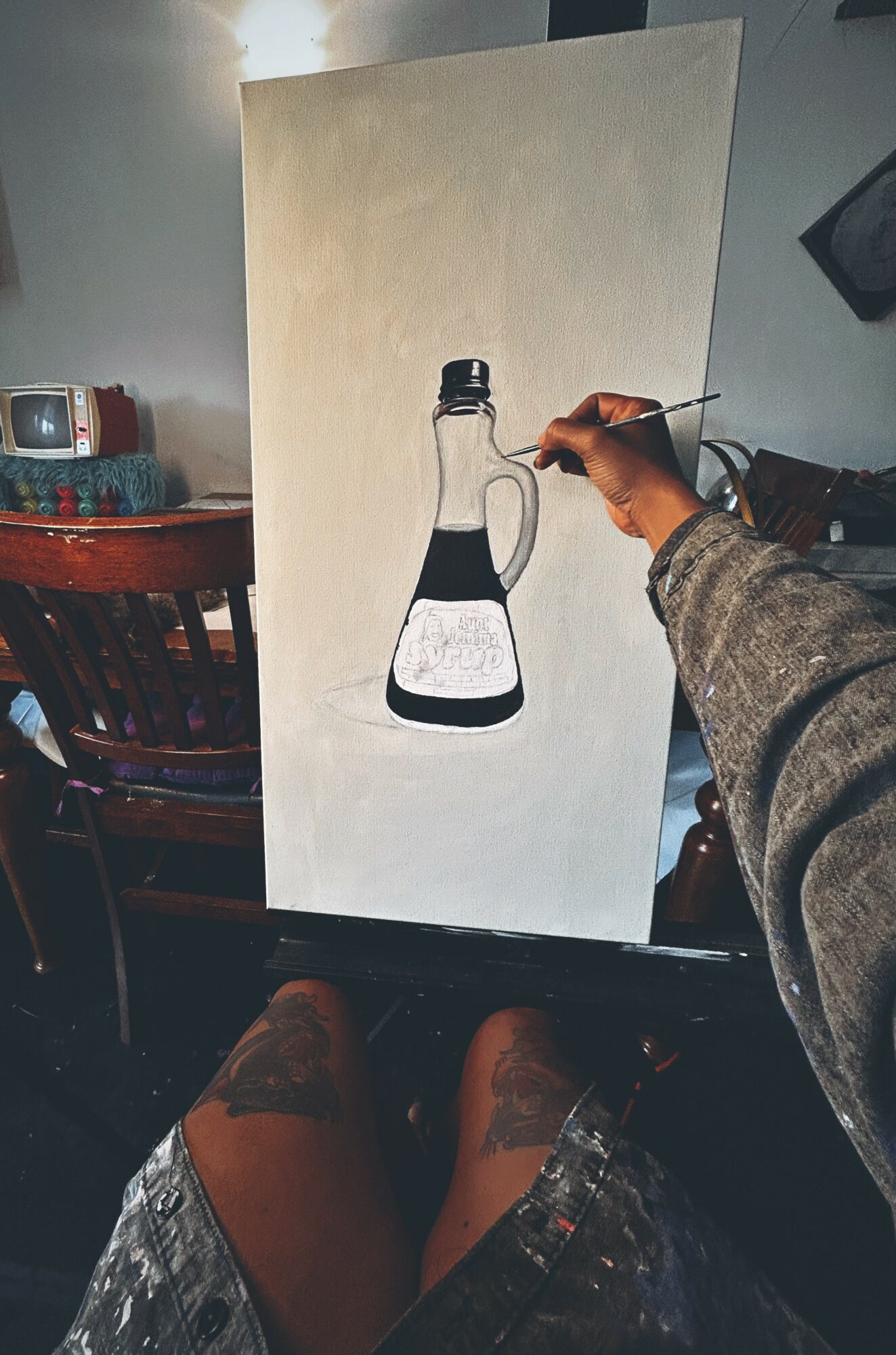Person painting a bottle with black and white liquid on a canvas, seated indoors with furniture visible.