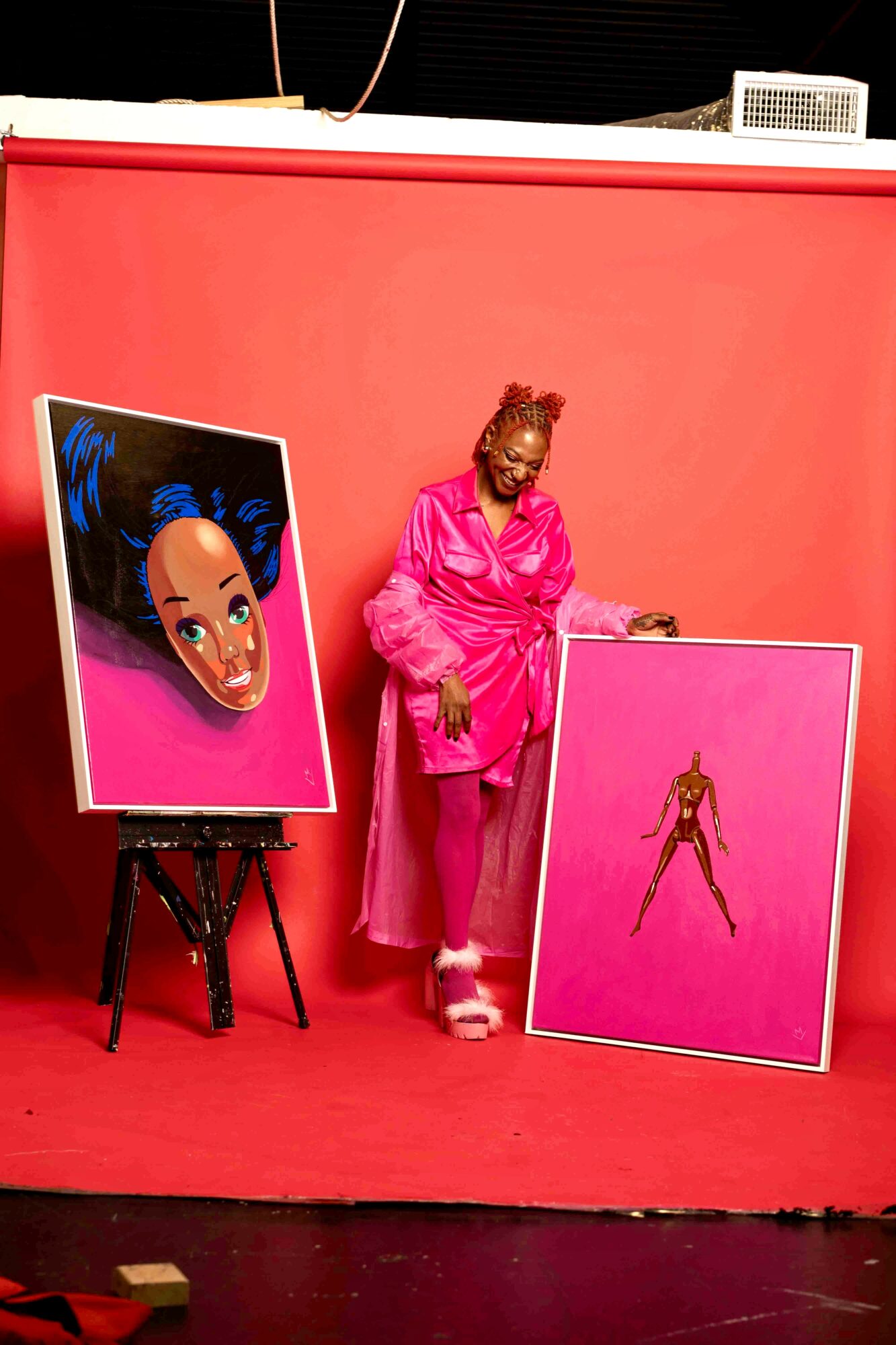 Woman in pink outfit standing next to two pink art pieces against a pink background.