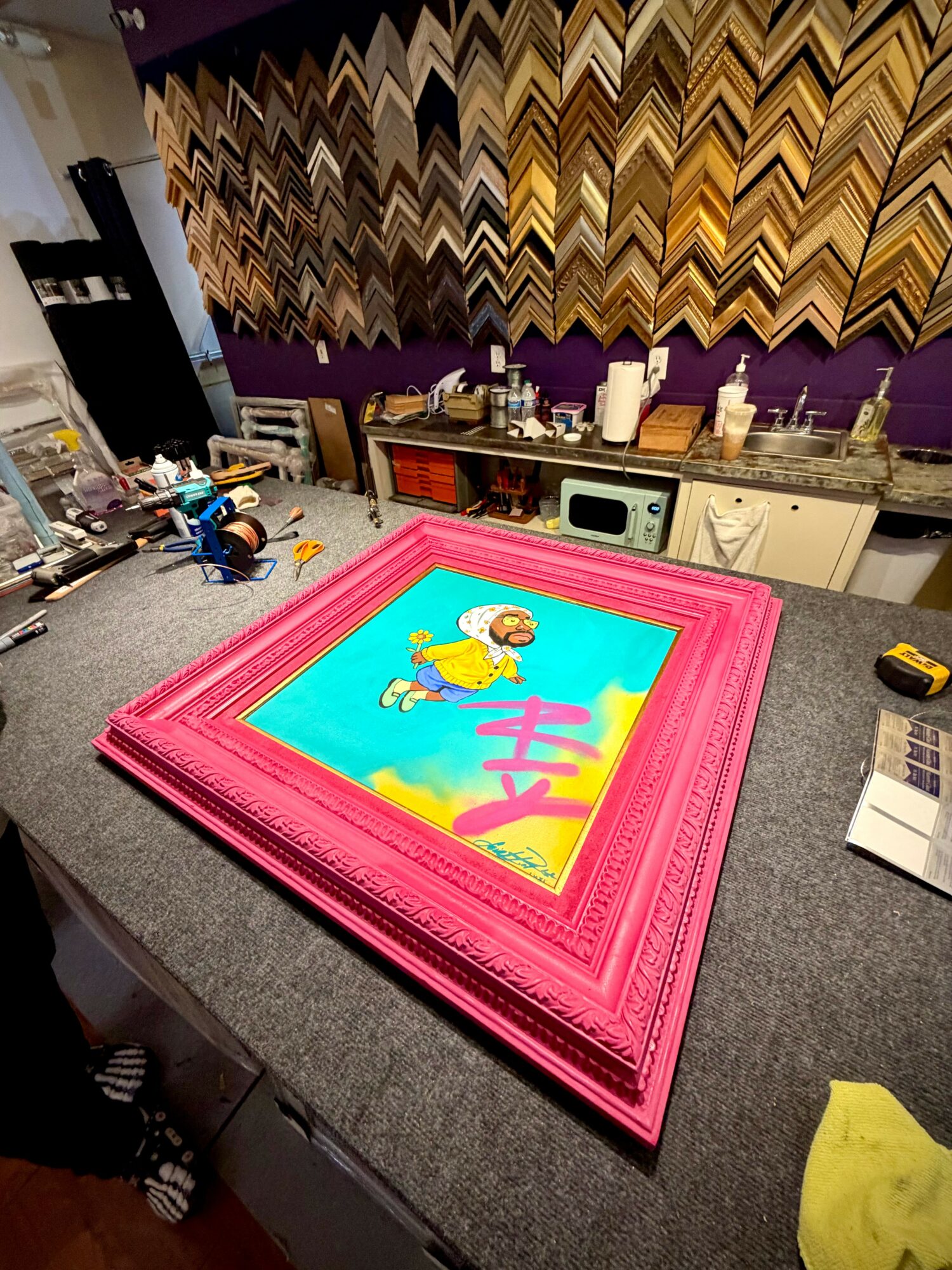 Colorful framed picture of a cartoon lion on a pink border, placed on a table in a room with patterned wall and kitchen.