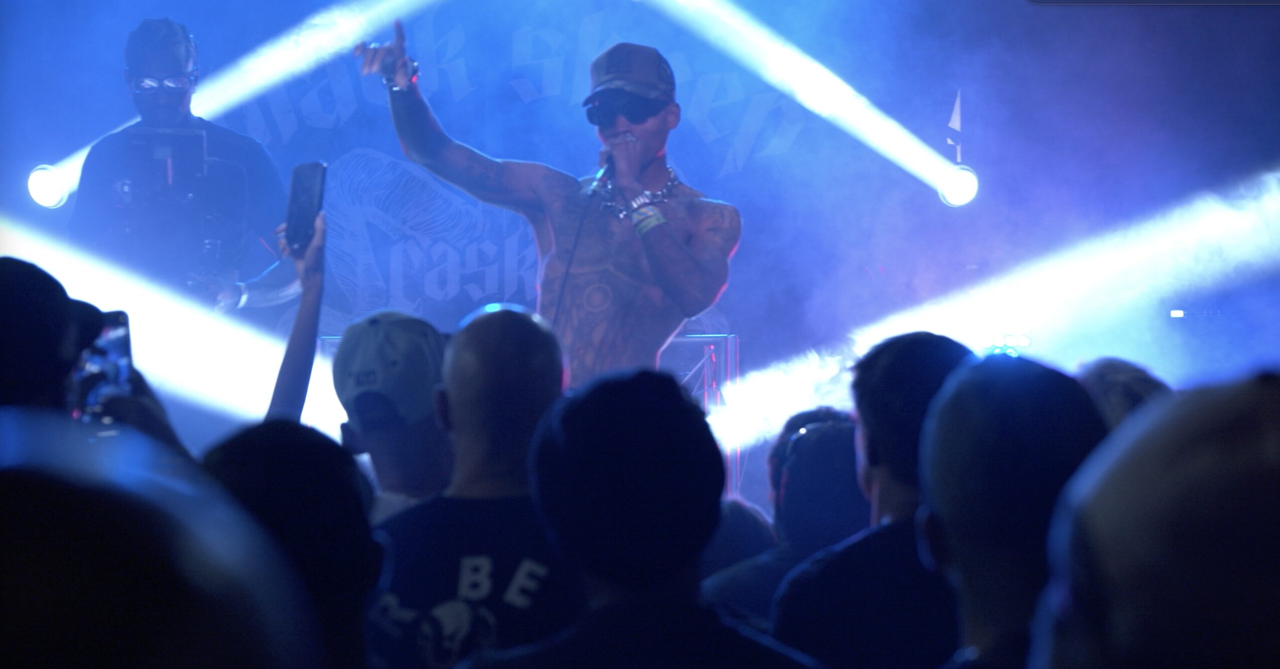 Performer on stage with microphone, wearing sunglasses and cap, audience watching in foreground, stage lights shining brightly.
