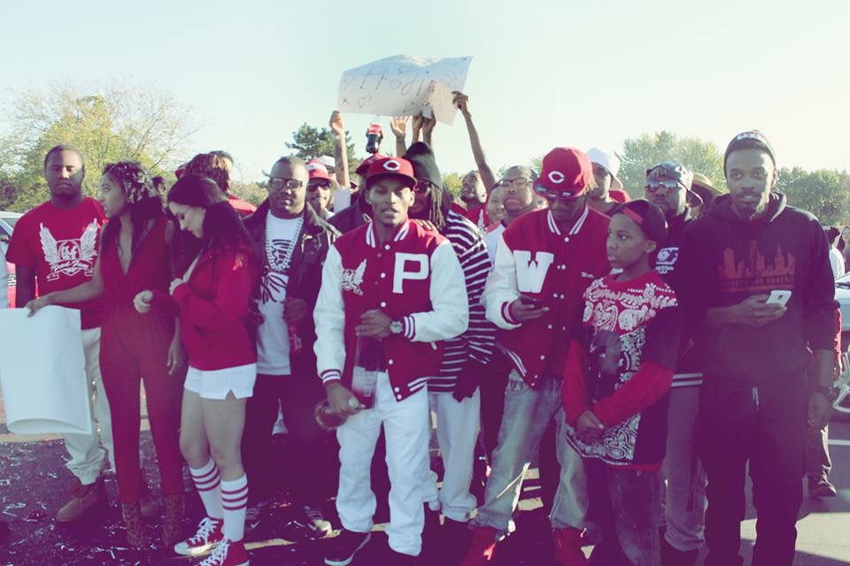 Group of people outdoors, some holding signs, wearing casual clothing and hats, standing together on a sunny day.