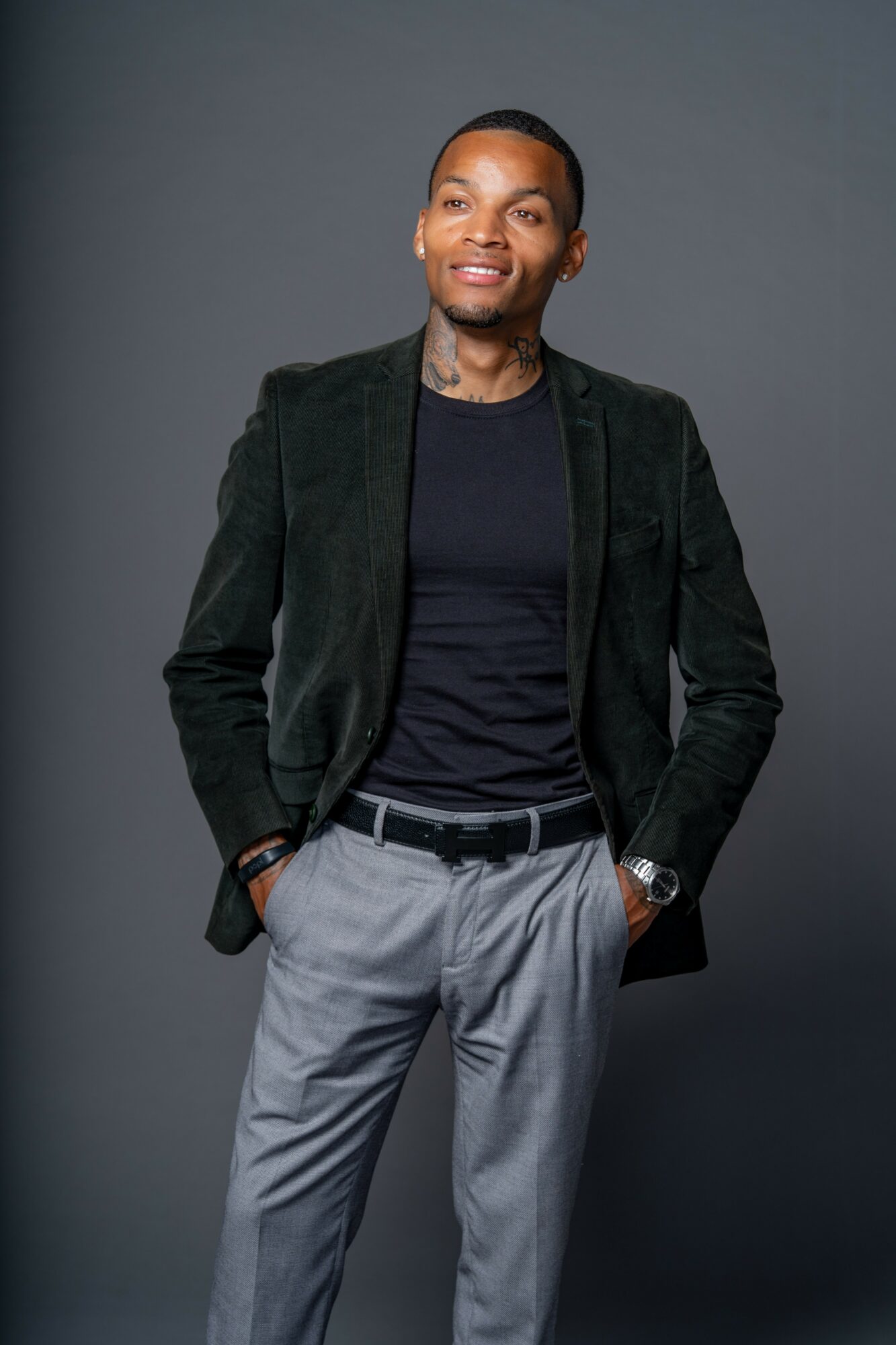 Man in black blazer and gray pants standing with hands in pockets, smiling, against gray background.